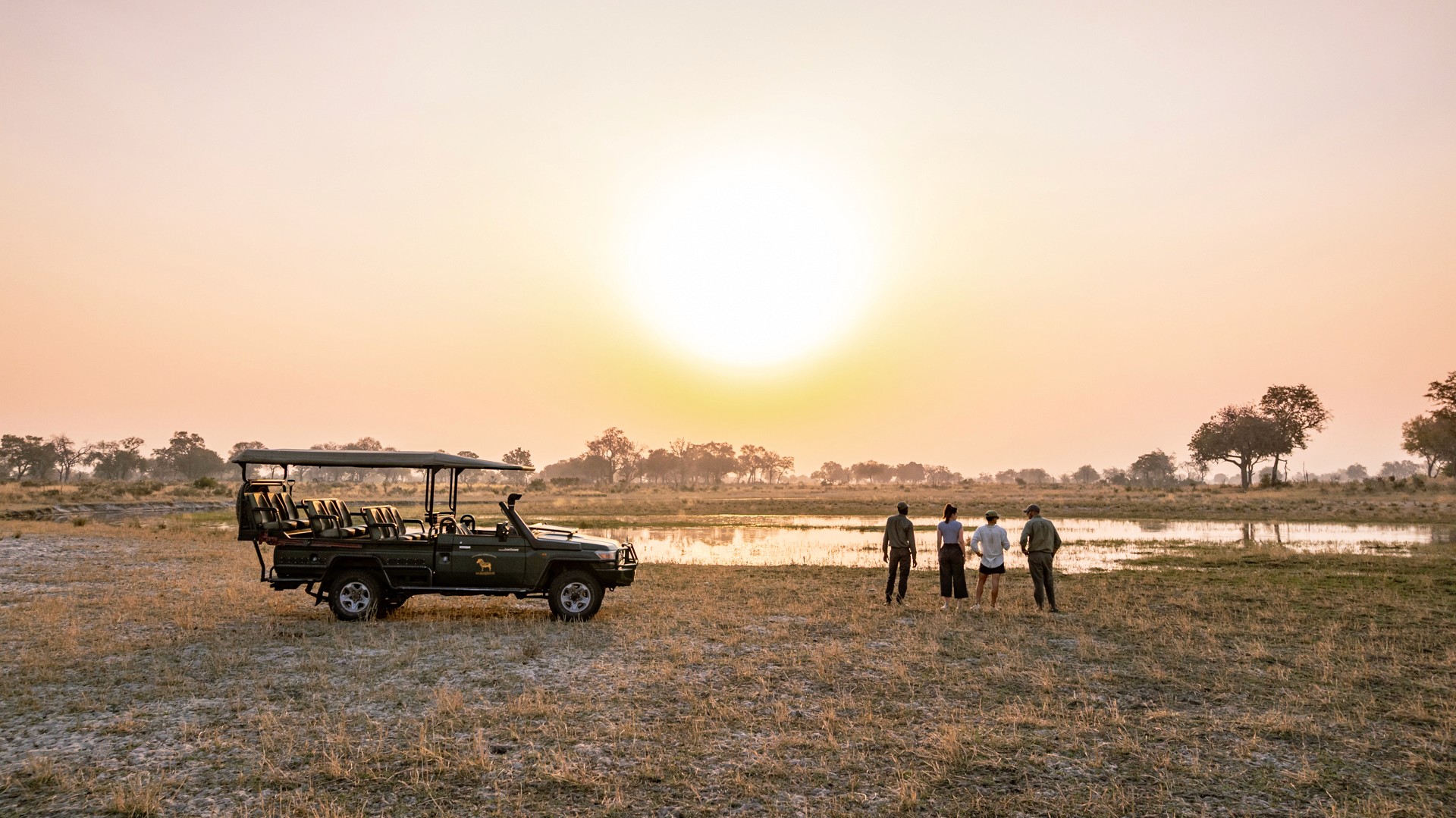 Kujwana Delta Ride, Okavango Delta, Botswana. Fyra personer på game drive blickar ut över solnedgången, med en safarijeep bredvid sig.