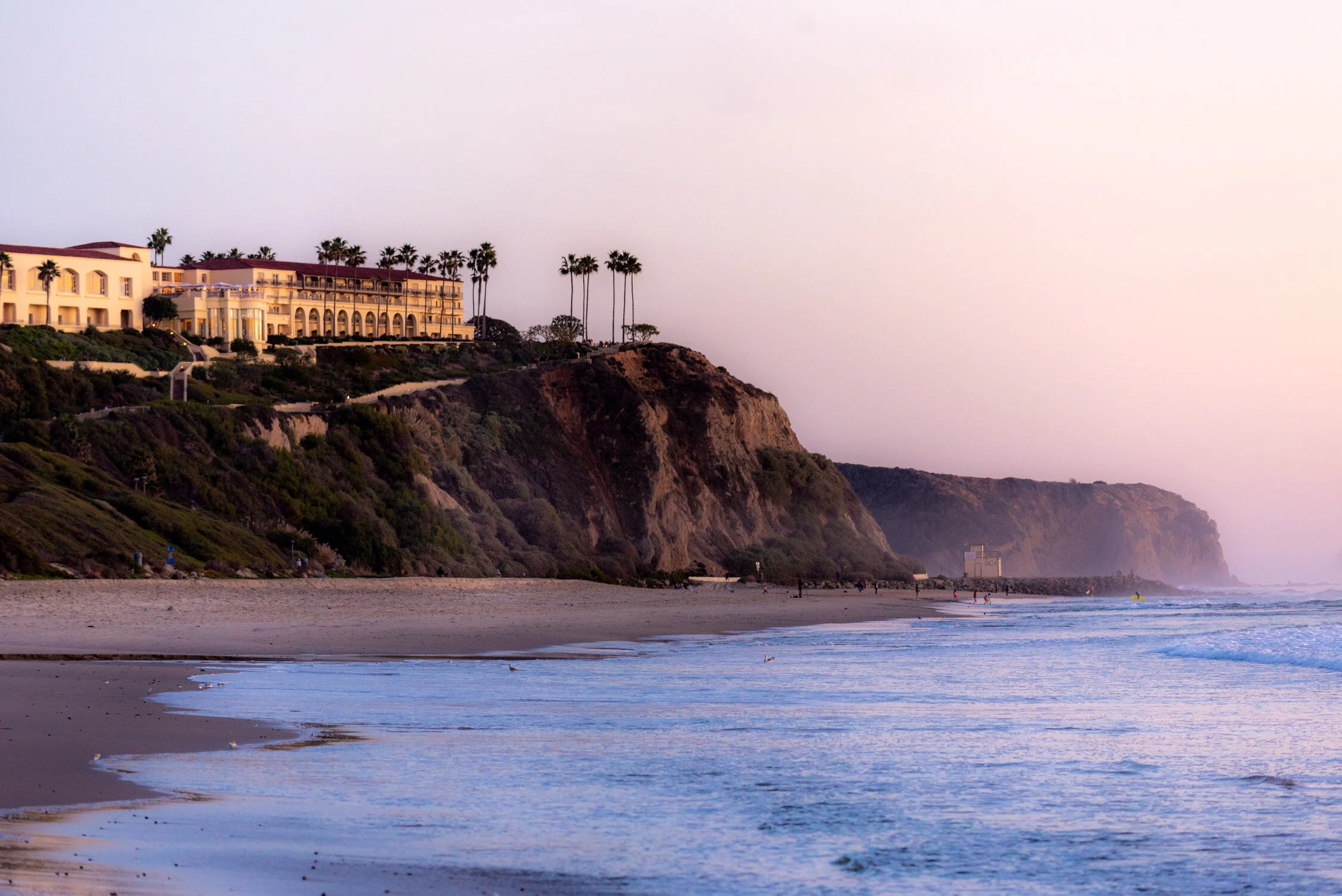 A coastal resort with a large tan building and palm trees sits atop a cliff overlooking a beach at dusk.
