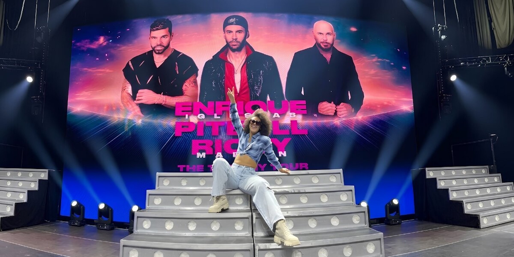 Woman posing on illuminated concert stage beneath Enrique Iglesias, Pitbull, and Ricky Martin tour backdrop.