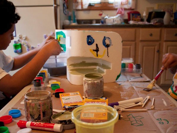 Two children are working on a craft project at a table covered in brown paper. The child on the left is using a paintbrush to apply green paint to a white cardboard box. The child on the right is using a paintbrush to paint a yellow smiley face on the box. Various craft supplies, including jars, paint tubes, and small containers, are scattered on the table. The background includes a kitchen counter with cabinets and a refrigerator.