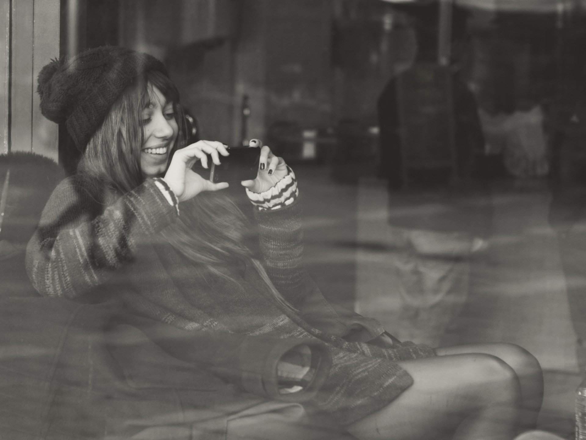 Artistic black-and-white street-style photograph showing a person in a knit hat and plaid coat using a smartphone behind a reflective window, the layered reflections adding depth and a candid, introspective mood.