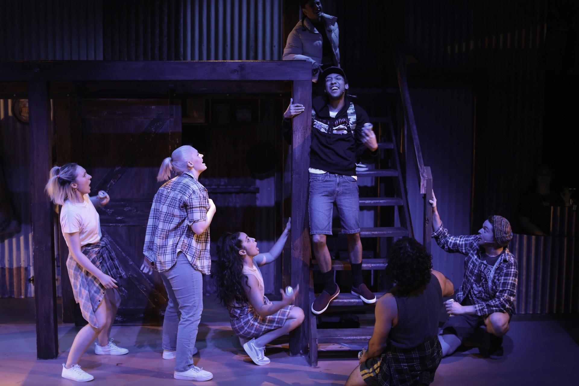 A group of young performers gather around a wooden platform on stage, animated and mid-scene. One performer stands on the lower steps of a staircase, laughing with a drink can in hand, while others reach toward him from below. The set is rustic, with corrugated metal walls and warm theatrical lighting.