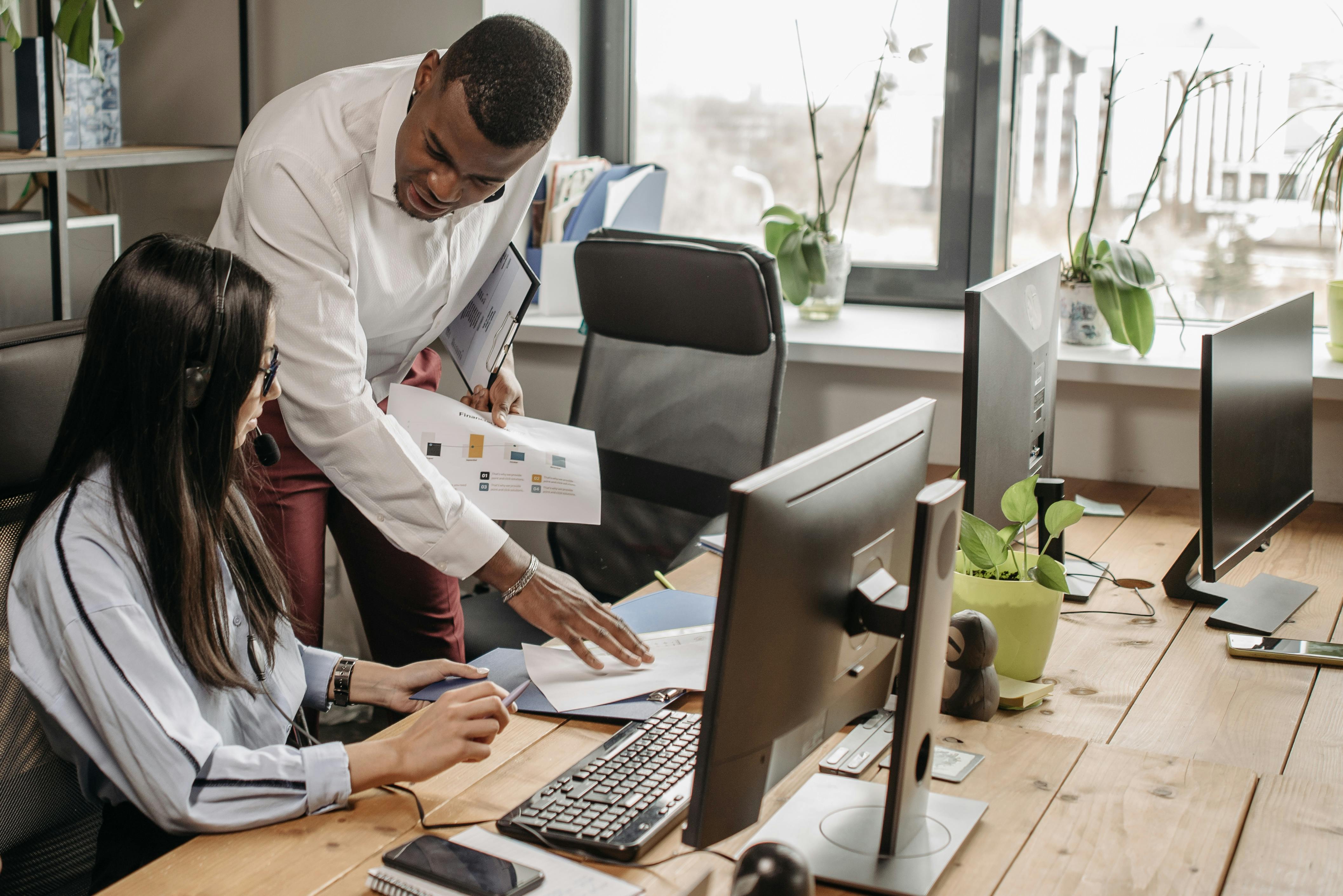 An image of two team members in an office space, sat at a desk. A taller male is leaning forward whilst stood up, looking at the female next to him as she works on something at the desk. She is paying attention to what he is saying.