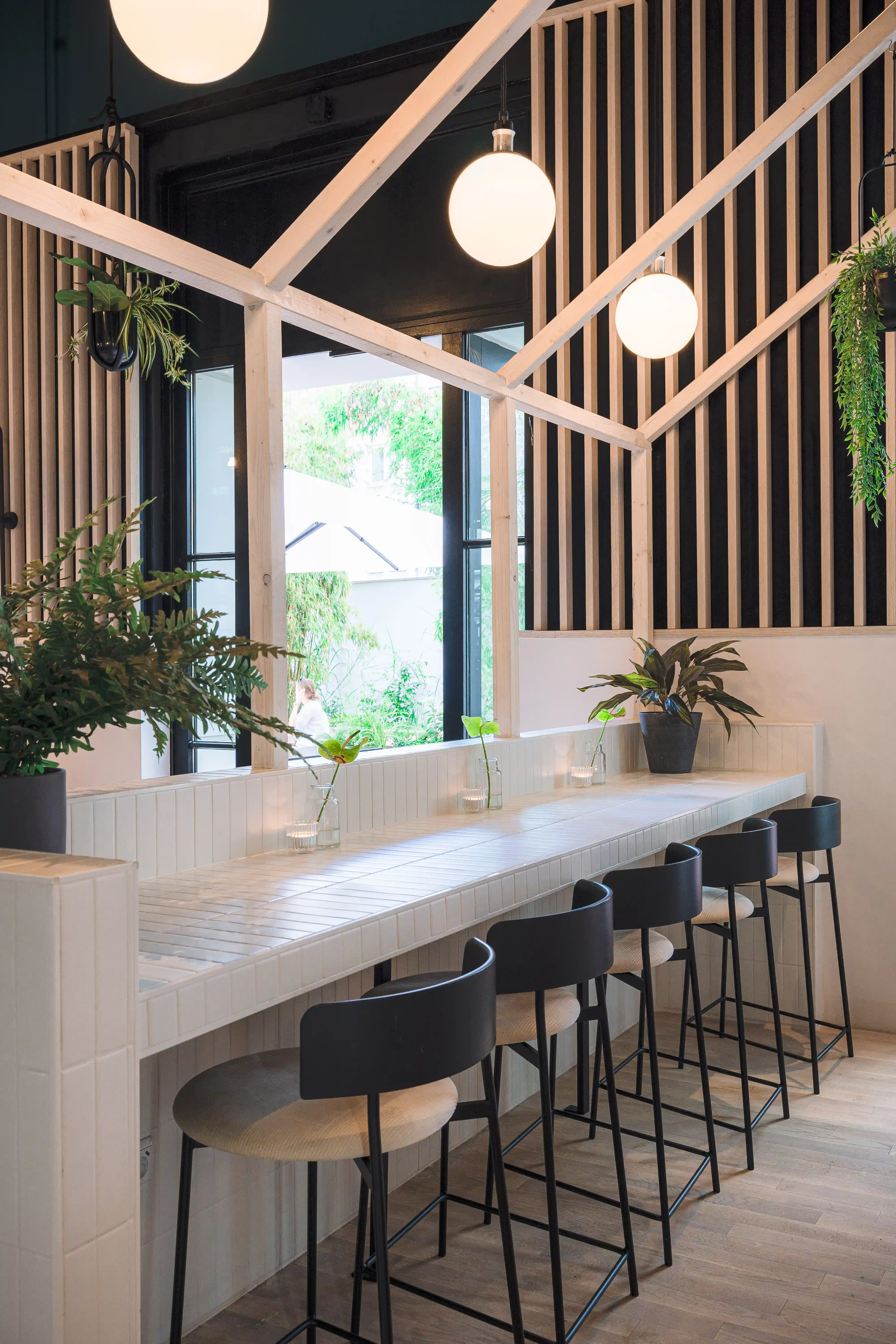 White tiled counter at LOKAL VIER with black bar stools, plants and tealights, framed by a white steel structure with globe lights and a view into the green courtyard.