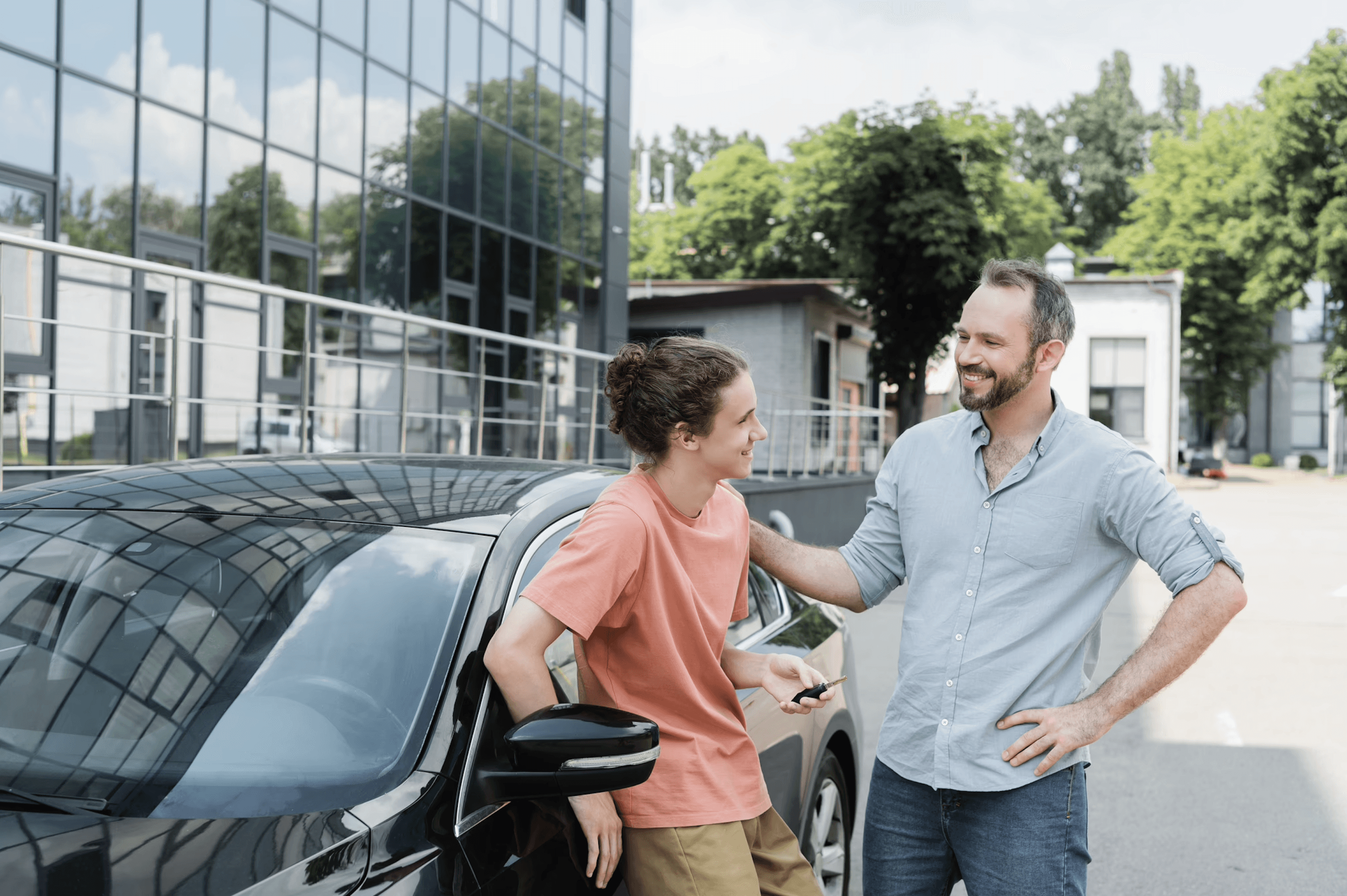 Smiling father and teenage son standing beside a black car, sharing a moment of joy as the son holds the car keys.