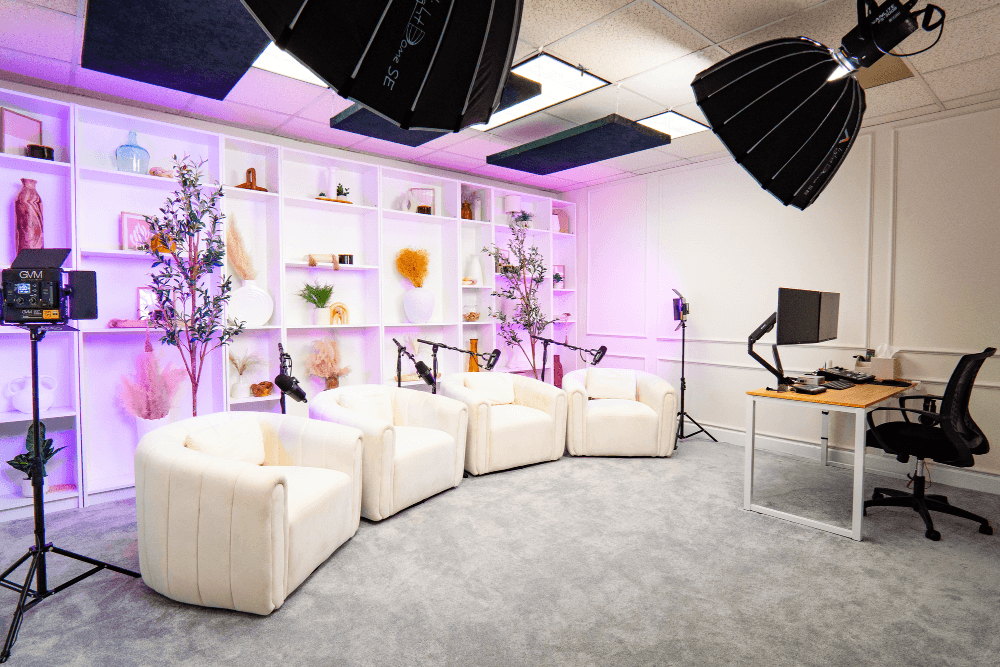 Book Studio B “Studio B featuring four white rounded chairs and a central ottoman on a light gray carpet, with a decorative shelf wall and soft pink-purple lighting creating a cozy, modern vibe.”