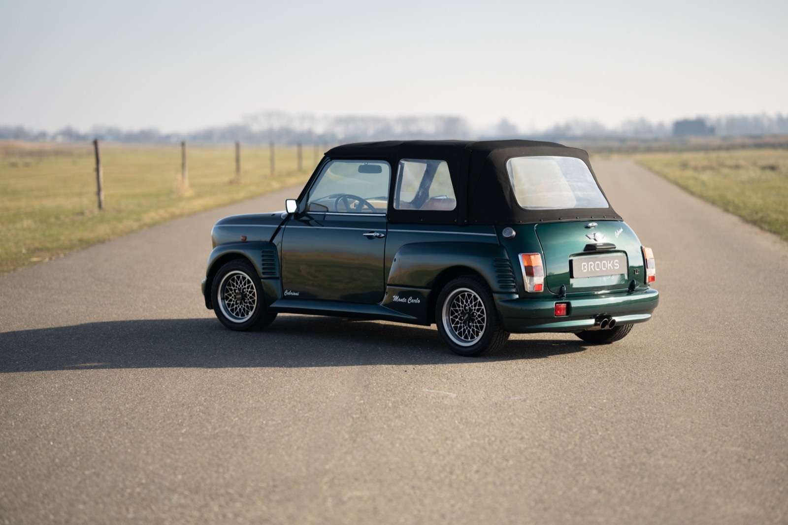 A classic car parked on a deserted road, surrounded by open fields under a clear blue sky.