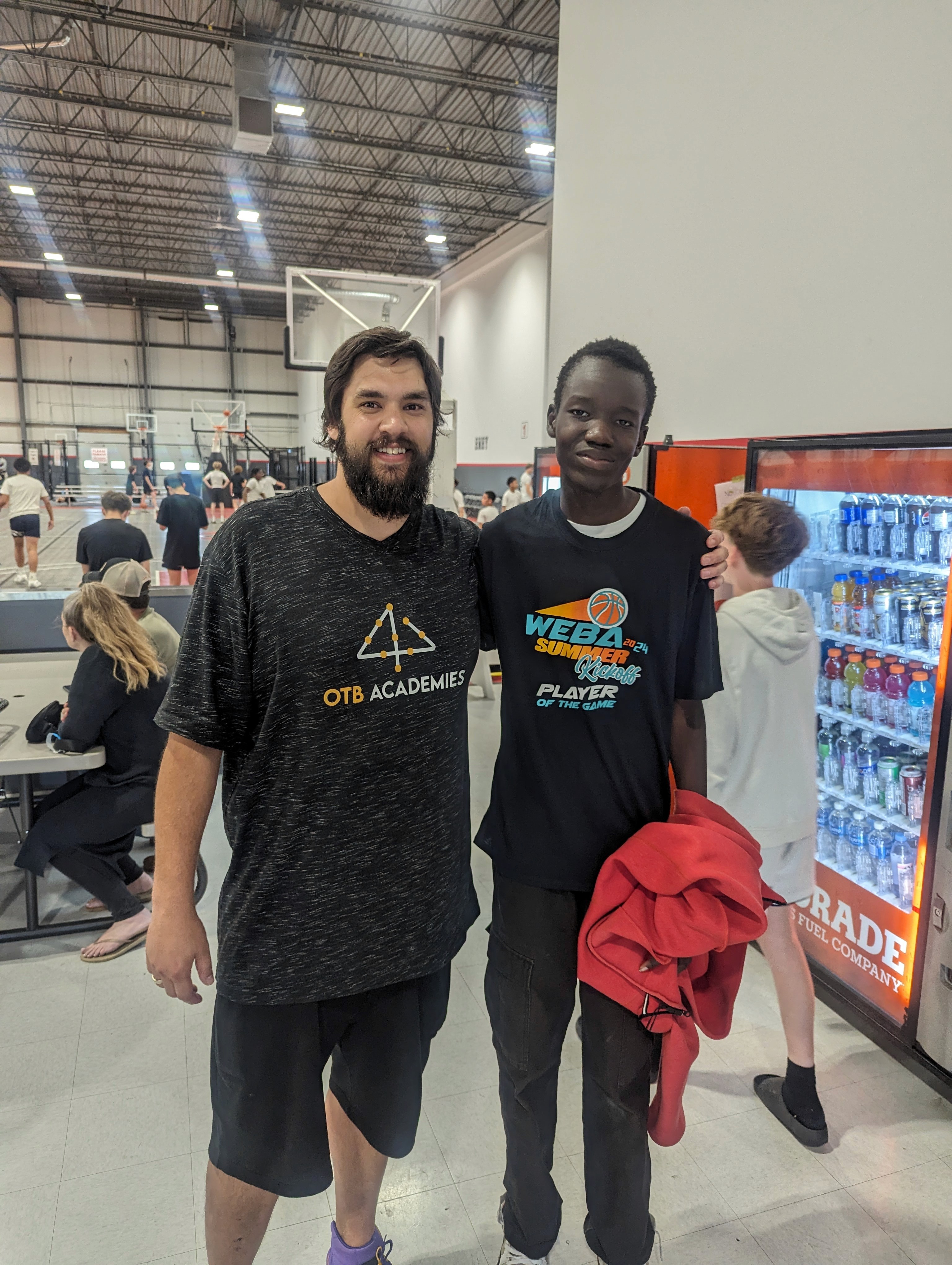 A youth basketball player in an orange jersey dribbling aggressively past a defender during a game, with a referee and spectators visible in the background on an indoor court.