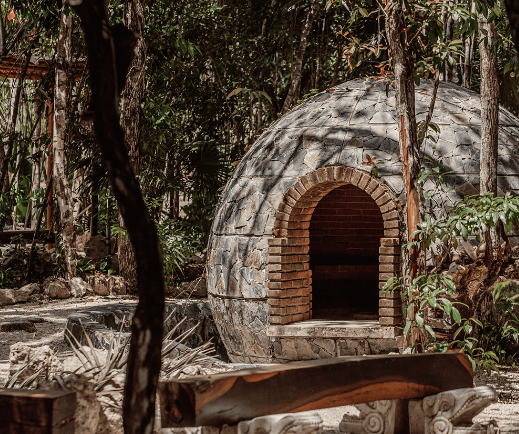 A family smiles & shares a meal at a rustic table outdoors amid a vineyard with a building in the background.