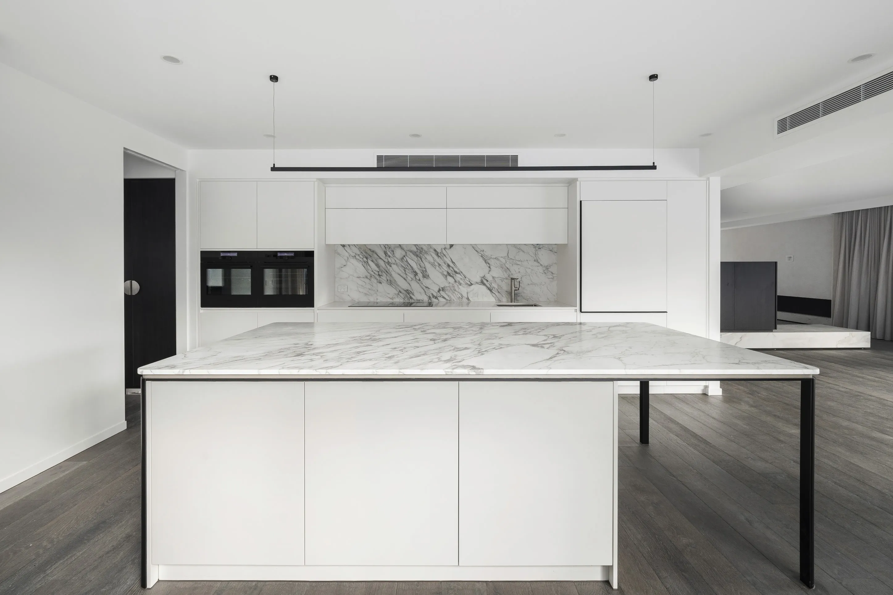 Kitchen viewed head-on with a large white marble island on black steel legs, marble splashback, white cabinetry, black wall ovens and dark timber floors