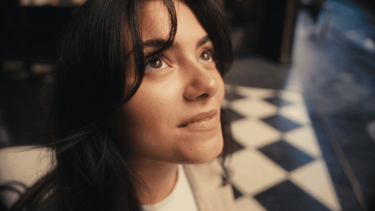 A woman looking upwards, standing on a checkered floor with soft lighting, wearing an elegant outfit