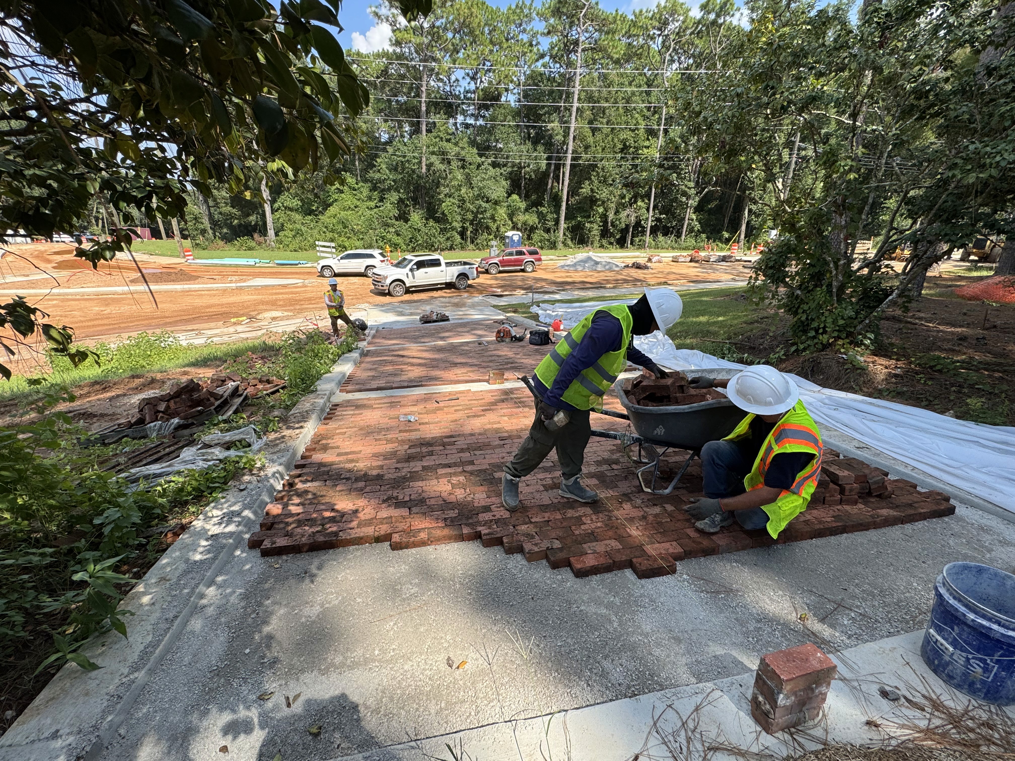 A group of 3–5 multiethnic professional landscape construction workers standing together on a sunny residential job site, smiling while holding tools like shovels, rakes, or a wheelbarrow. They are wearing clean branded work shirts or vests, work gloves, and hats. The background shows an active landscape project with sod, mulch, stonework, and garden beds in progress. Clear blue sky, bright natural lighting, friendly expressions, realistic proportions, and professional photo composition.
