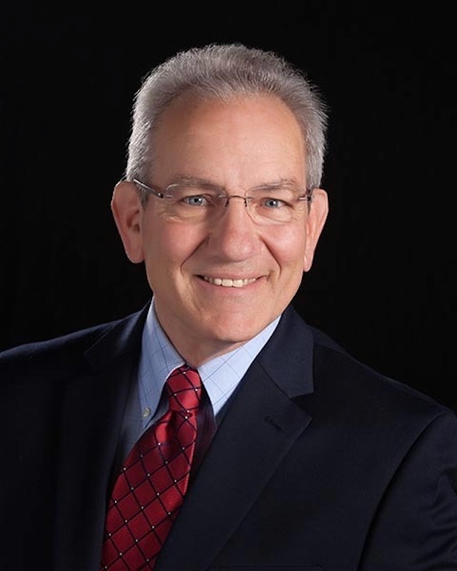 A man with gray hair and glasses smiles confidently against a black background. He wears a navy suit, light blue shirt, and red patterned tie.