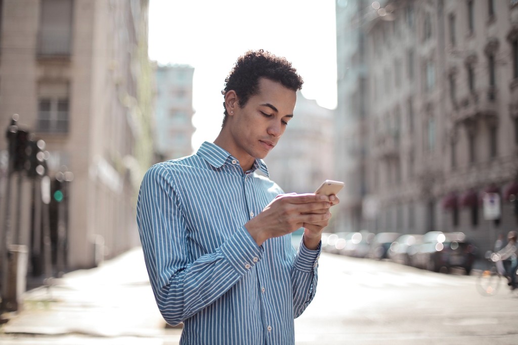 Man texting on smartphone in urban street, wearing blue striped shirt.