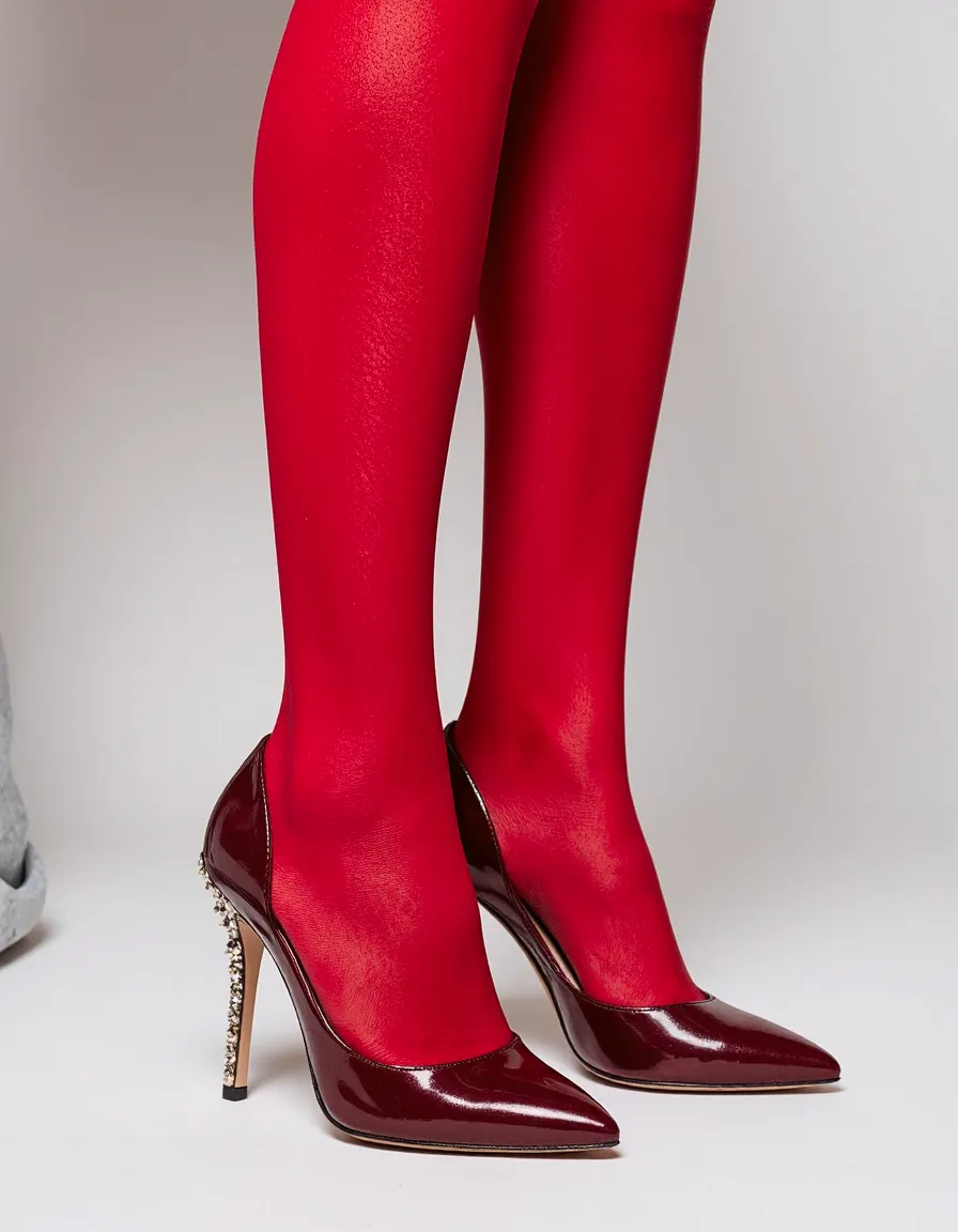 Burgundy patent leather stilettos with embellished heels worn with bright red tights against a neutral background