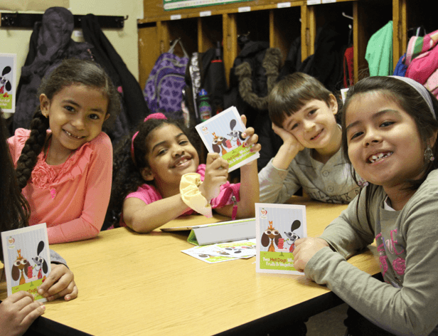 Four young girls are seated around a light brown table in a classroom setting. The girl on the far left wears a pink long-sleeved shirt and has long dark hair styled in two braids. Next to her, a girl in a pink short-sleeved shirt smiles at the camera. The third girl wears a gray long-sleeved shirt and has short brown hair resting on her hand. The girl on the far right wears a gray long-sleeved shirt with a light-colored collar and smiles at the camera. Each girl holds a colorful card featuring a cartoon dog. Behind them, shelves hold backpacks and coats, and a purple backpack is visible.