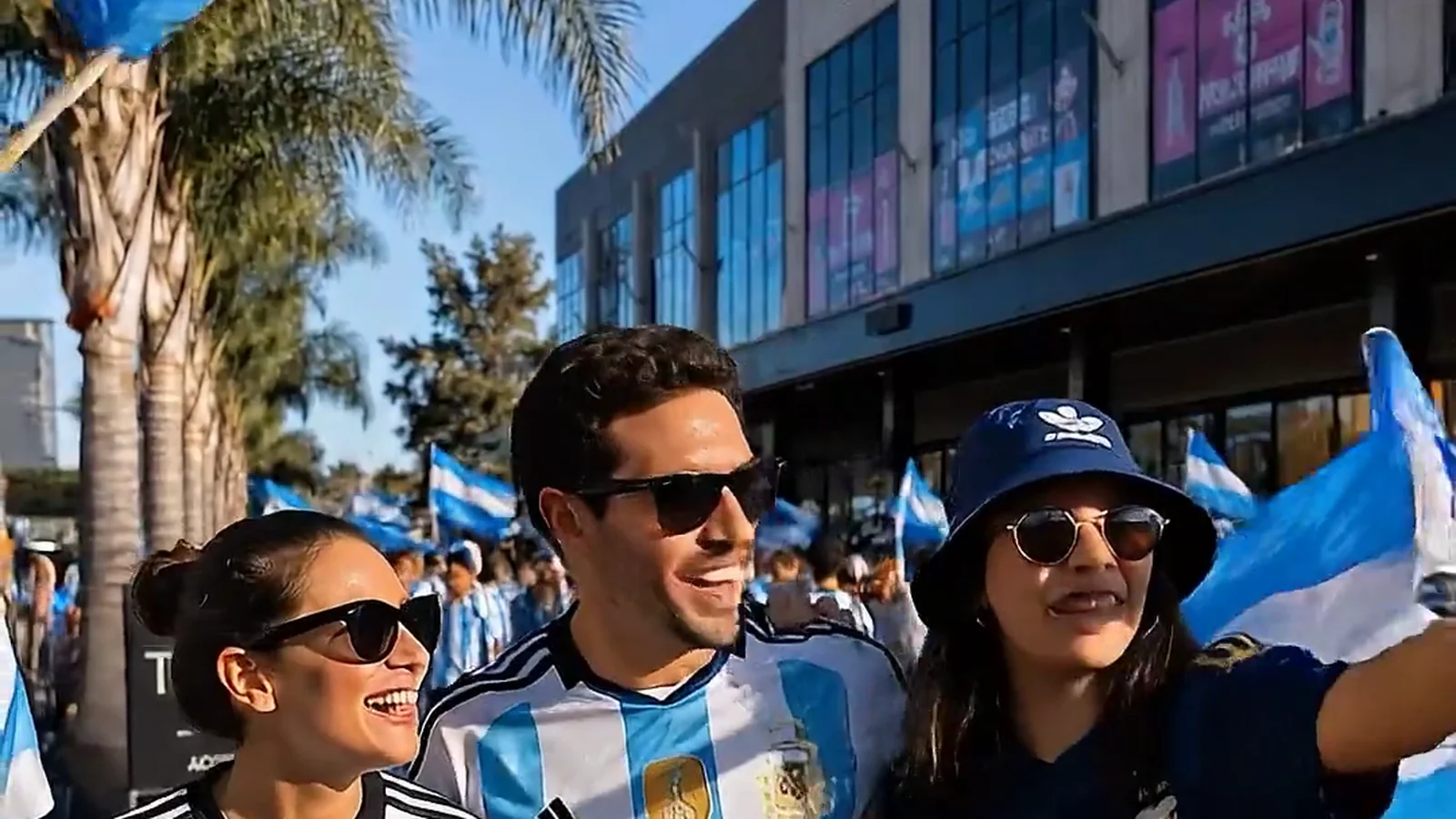 Fans proudly wearing Argentina jerseys and waving national flags celebrate outside Toscas Shopping on a sunlit afternoon rally.