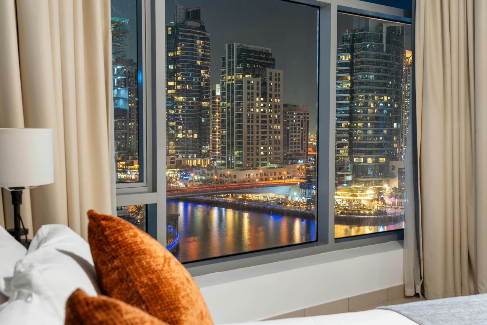 A bedroom with a white bed, orange pillows, and large windows overlooking the Dubai Marina skyline and waterfront at night.