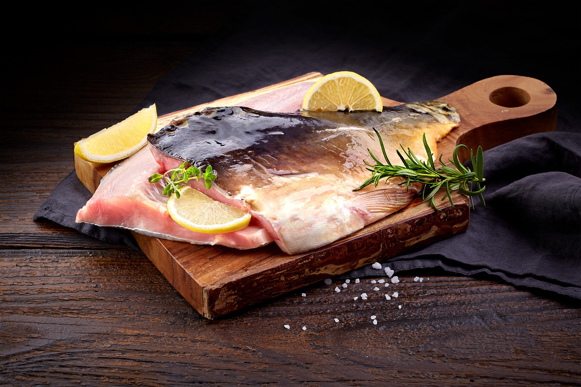 A fresh fish carp fillet on a wooden cutting board garnished with lemon slices, rosemary, and coarse sea salt, set against a dark rustic wooden background.
