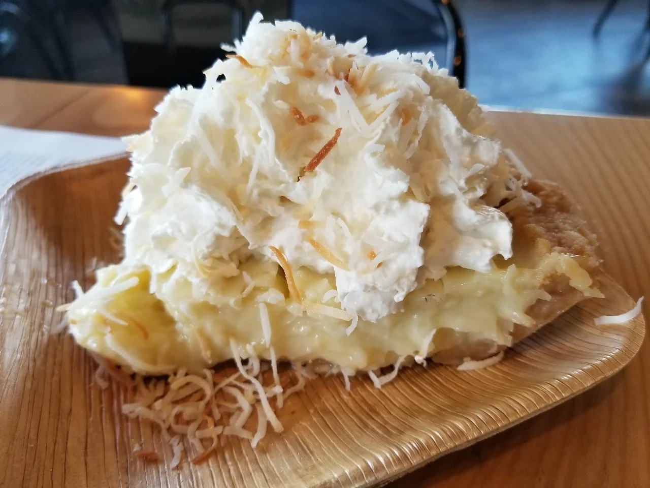 A large slice of coconut cream pie with a thick layer of pale yellow custard, a cookie crust, and a massive topping of white whipped cream and toasted coconut shavings.