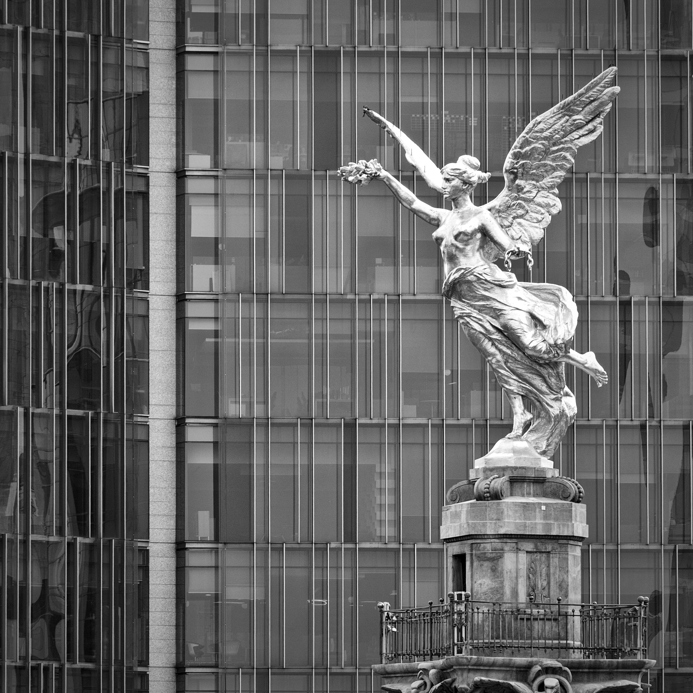 Black and white photograph of the Ángel de la Independencia statue in Mexico City, with a modern glass building in the background.