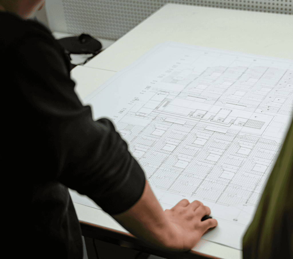 Detailaufnahme von zwei Mitarbeitenden der kba Architekten und Ingenieure GmbH, die über großformatige technische Bauzeichnungen gebeugt sind und Grundrisse analysieren; Fokus auf den Plan und die Hände über dem Tisch.