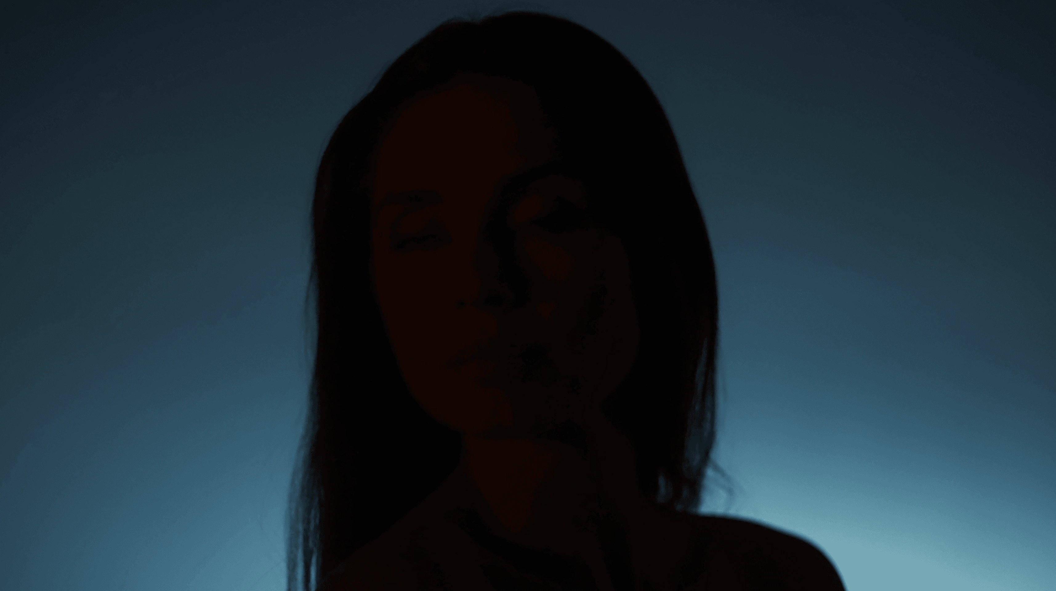 In a softly lit, atmospheric image, a silhouette of a person with long hair stands against a gradient blue background, evoking a sense of mystery and introspection.