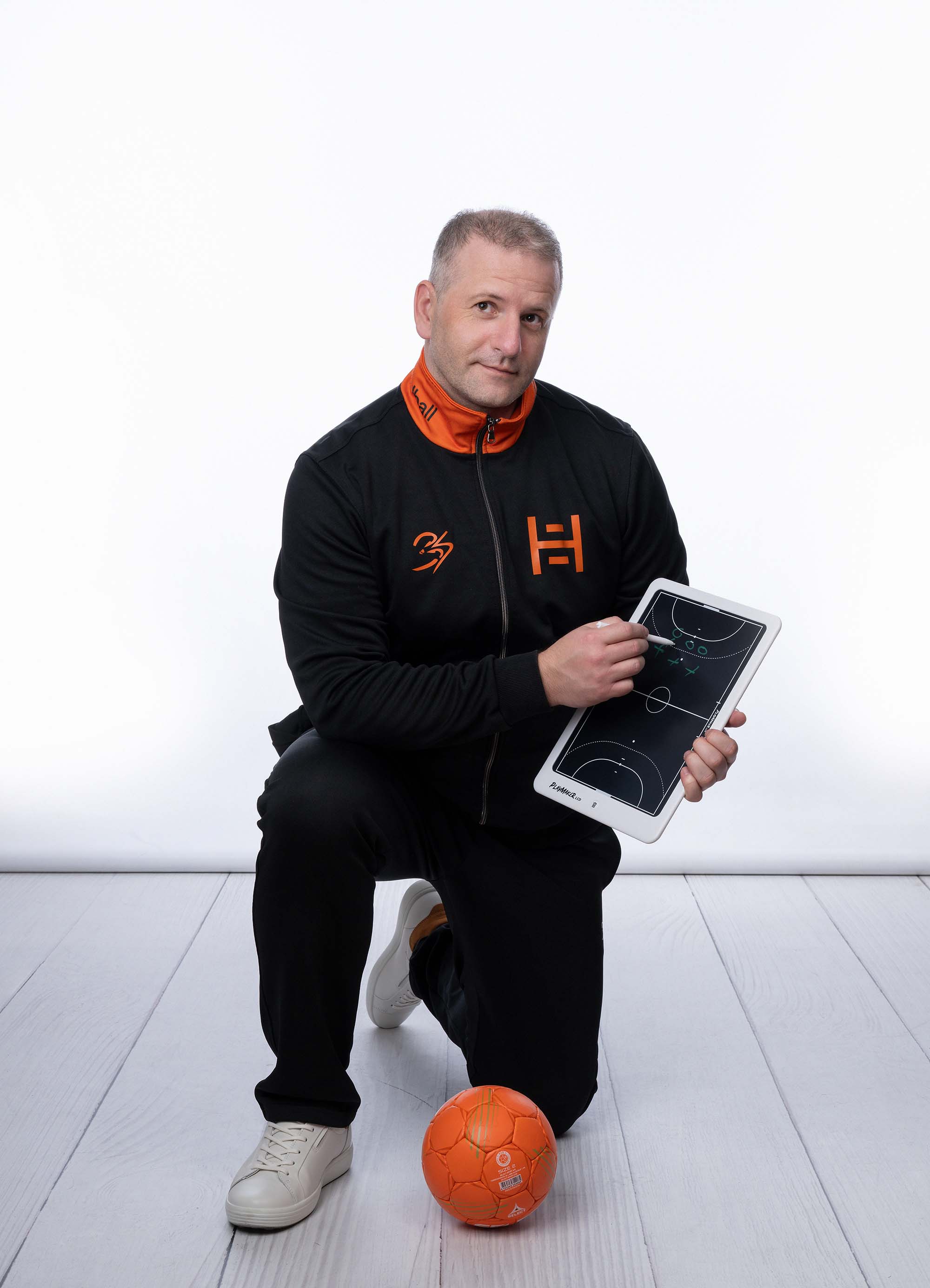 A handball coach kneeling beside an orange ball, drawing up a play on a tactical board while wearing a black and orange tracksuit.