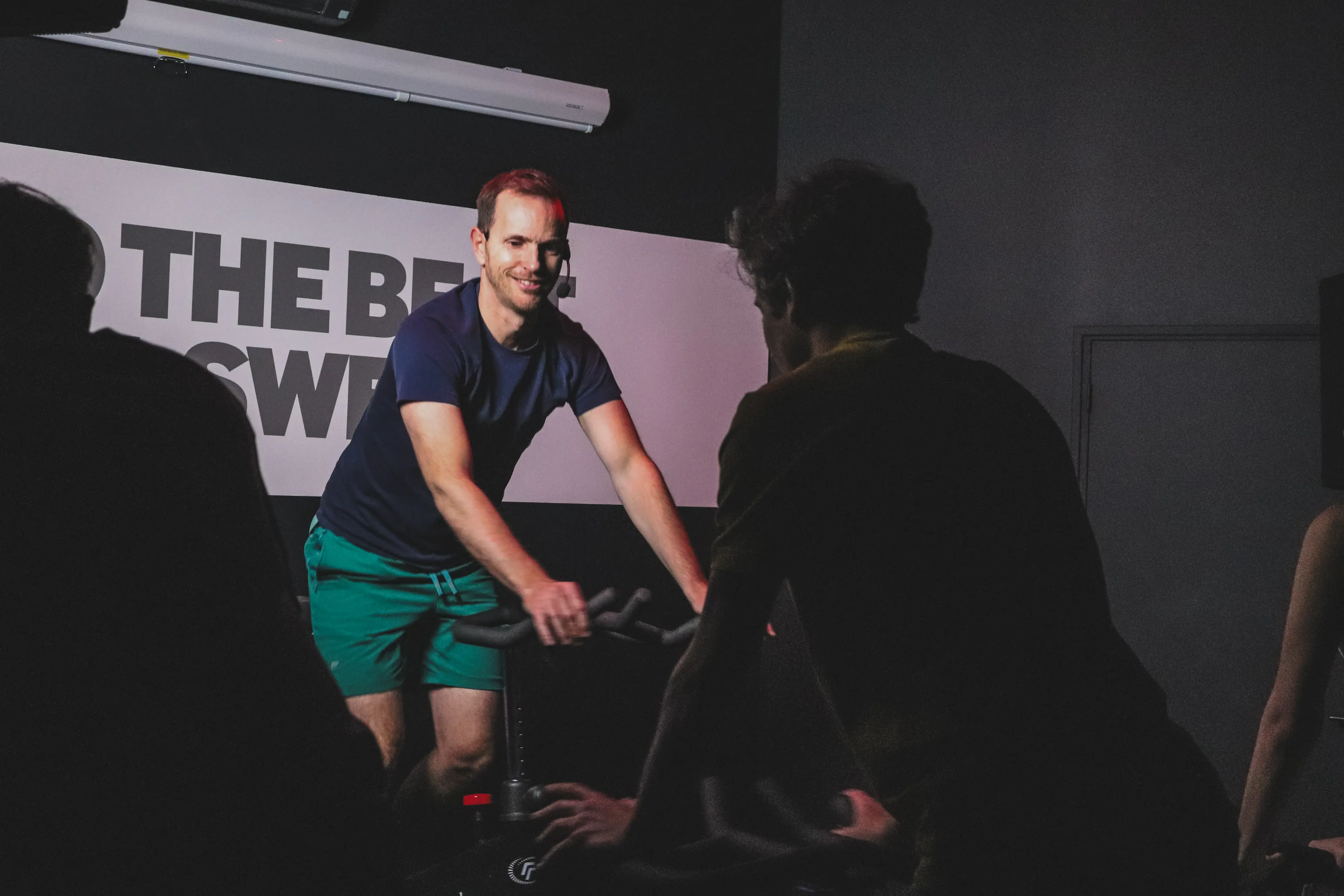 Coach guidant un cours de cycling en studio sombre, souriant face aux participants sur leurs vélos