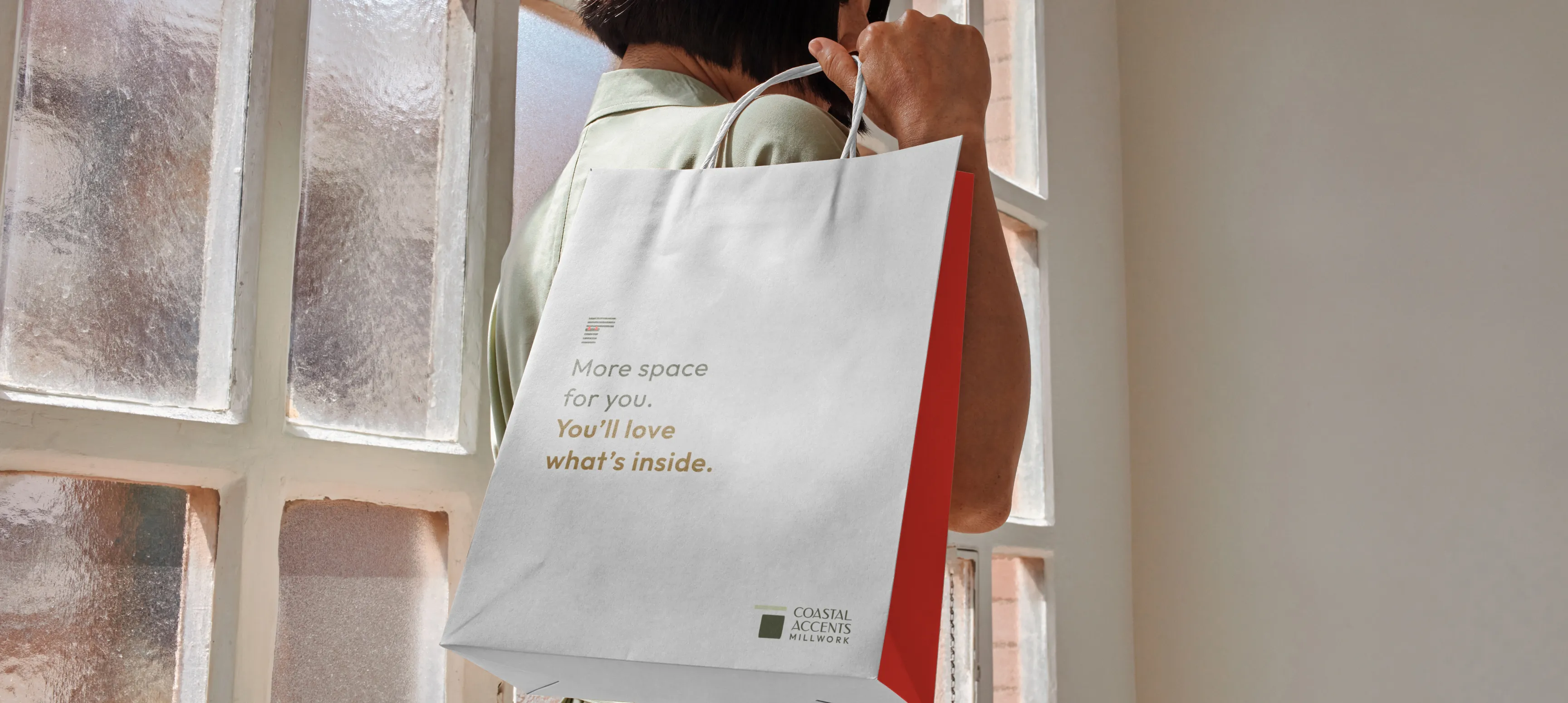 A woman standing in front of a window holding a paper retail bag that says "More space for you. You'll love what's inside" with a joinery element, the Coastal Accents logo, and red sides.