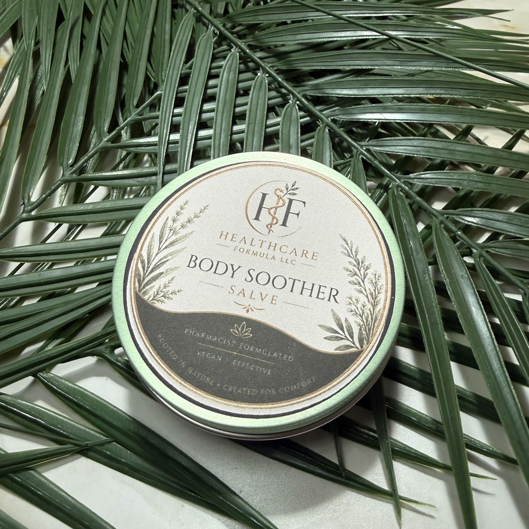 Healthcare Formula Body Soother Salve container displayed among dark green botanical leaves.