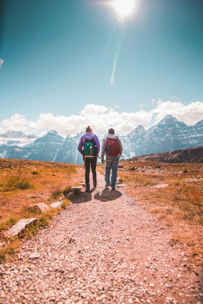 Two young backpackers hiking towards the snow-capped Canadian Rocky Mountains on a sunny day, representing international travelers arriving on an IEC Working Holiday visa ready to start their Canadian adventure.