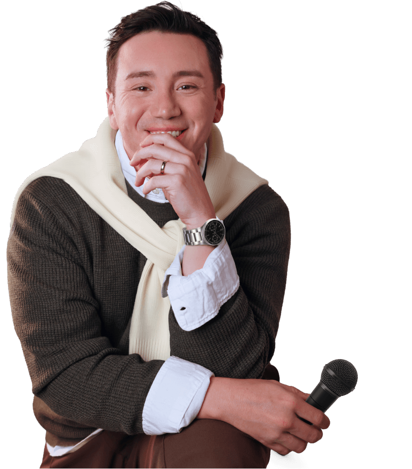 Retrato de Gianluca Peroti, sentado y sonriente con una mano en el rostro y otra con un micrófono, con un sweater amarrado al cuello manga larga, sobre un fondo tropical e incongruente con lo que lleva.