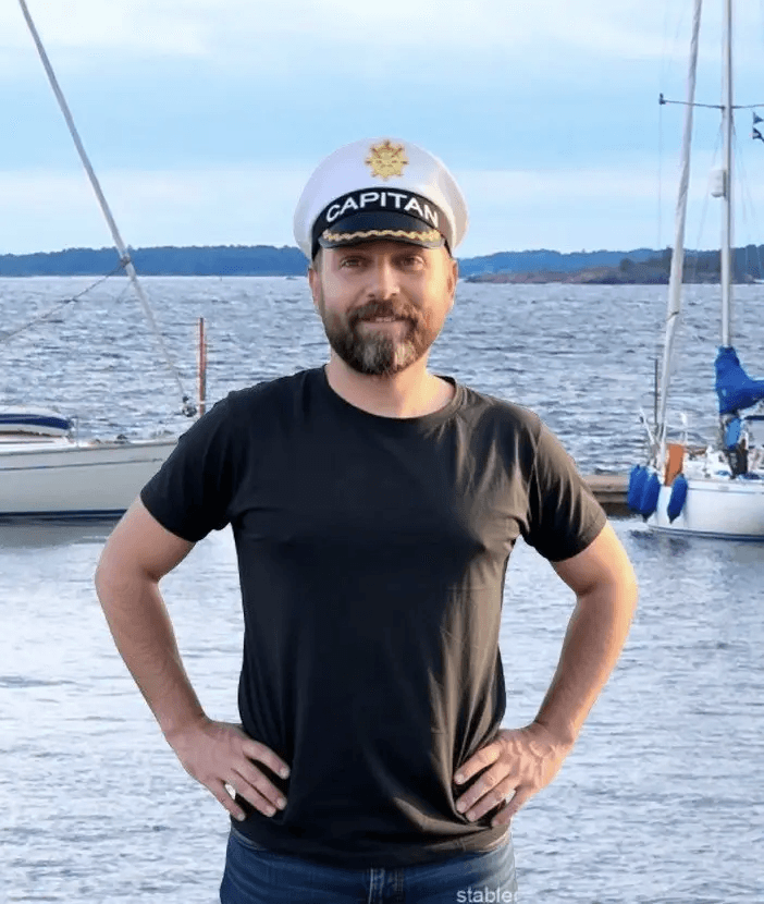 Skipper Malte mit weißer Kapitänsmütze und schwarzem Shirt vor ankernden Segelyachten am Wasser
