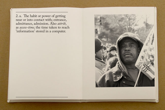 An open book displays two pages. The left page contains text defining "habit or power of getting near or into contact with entrance, admittance, admission. Also attrib, as access-time, the time taken to reach 'information' stored in a computer." The right page features a black and white photograph of a man in a hooded jacket holding a sign. The sign includes text that is partially visible but unreadable. The background of the photograph is out of focus and shows trees and a light-colored sky. The book rests on a light brown surface.
