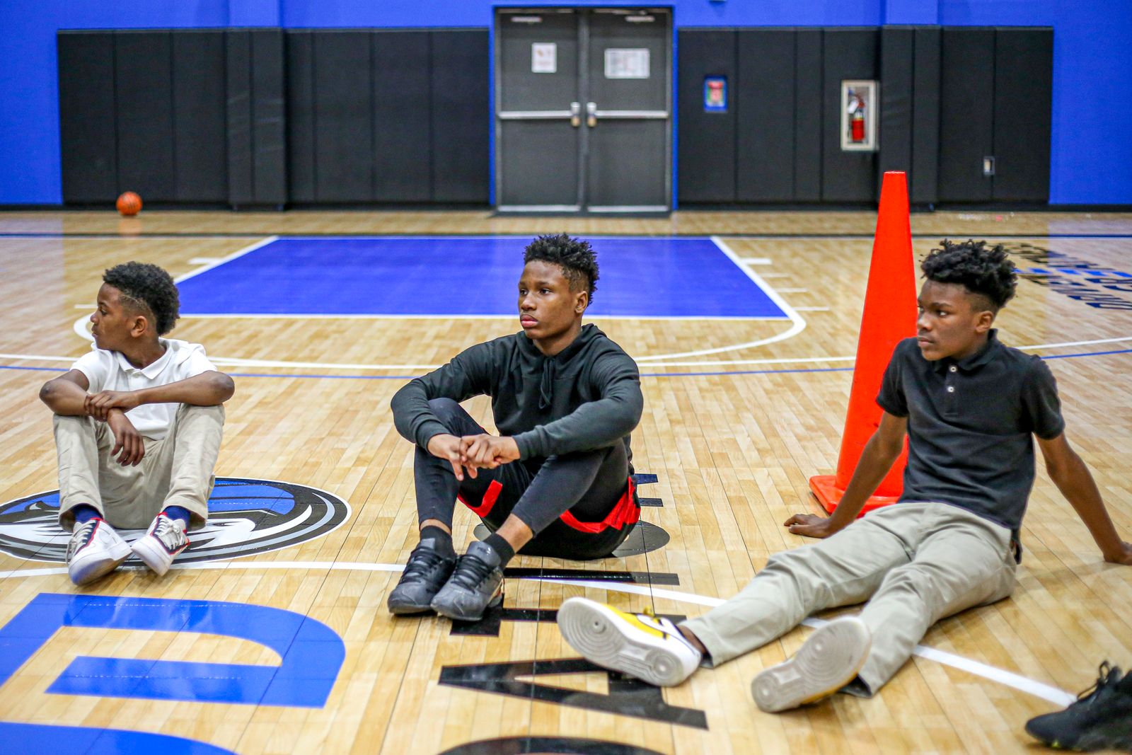 Three boys sitting on a basketball court during a break, with focus and contemplation on their faces.