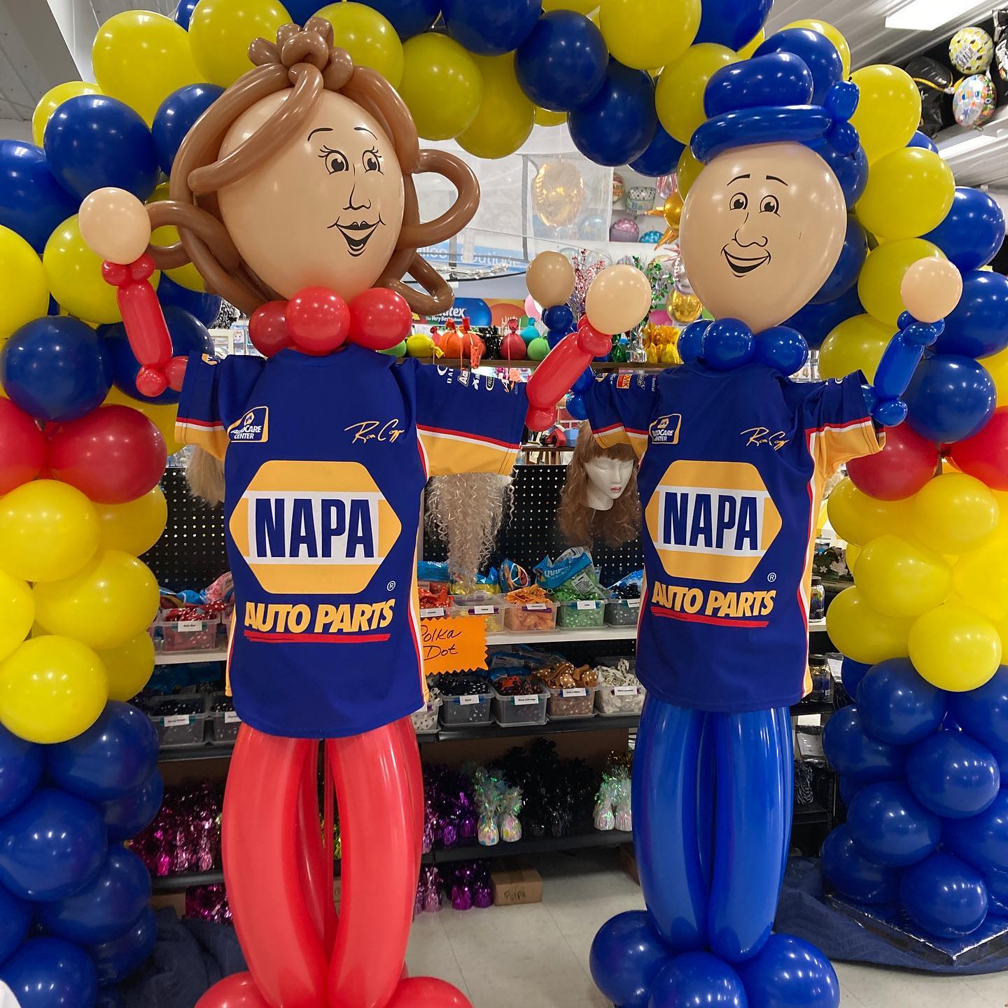 Balloon sculptures of smiling figures wearing NAPA Auto Parts jerseys stand under an arch made of blue and yellow balloons in a store filled with various party supplies.