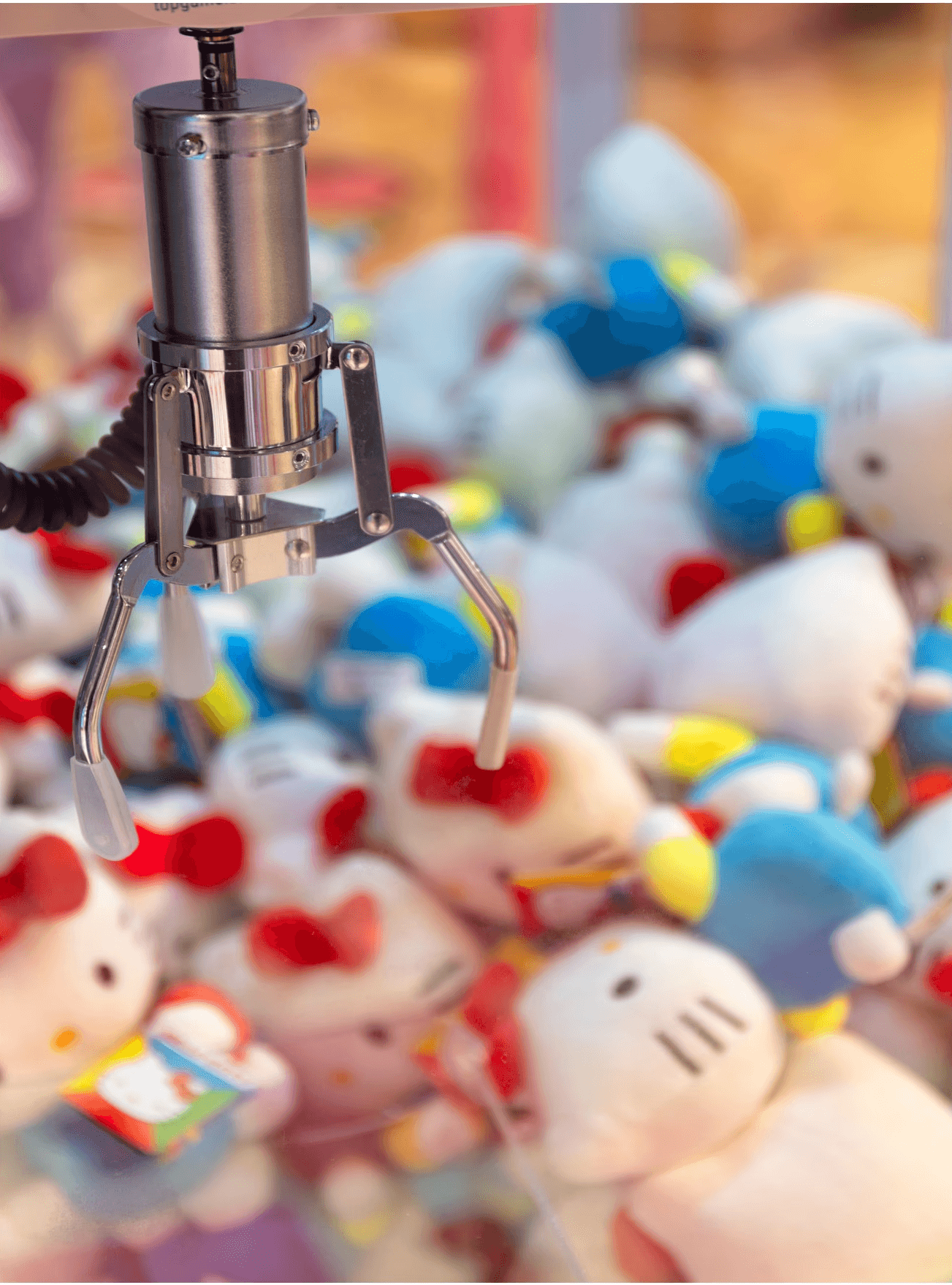 Gros plan sur la pince métallique d'une machine à gagner, au-dessus de peluches Hello Kitty colorées.