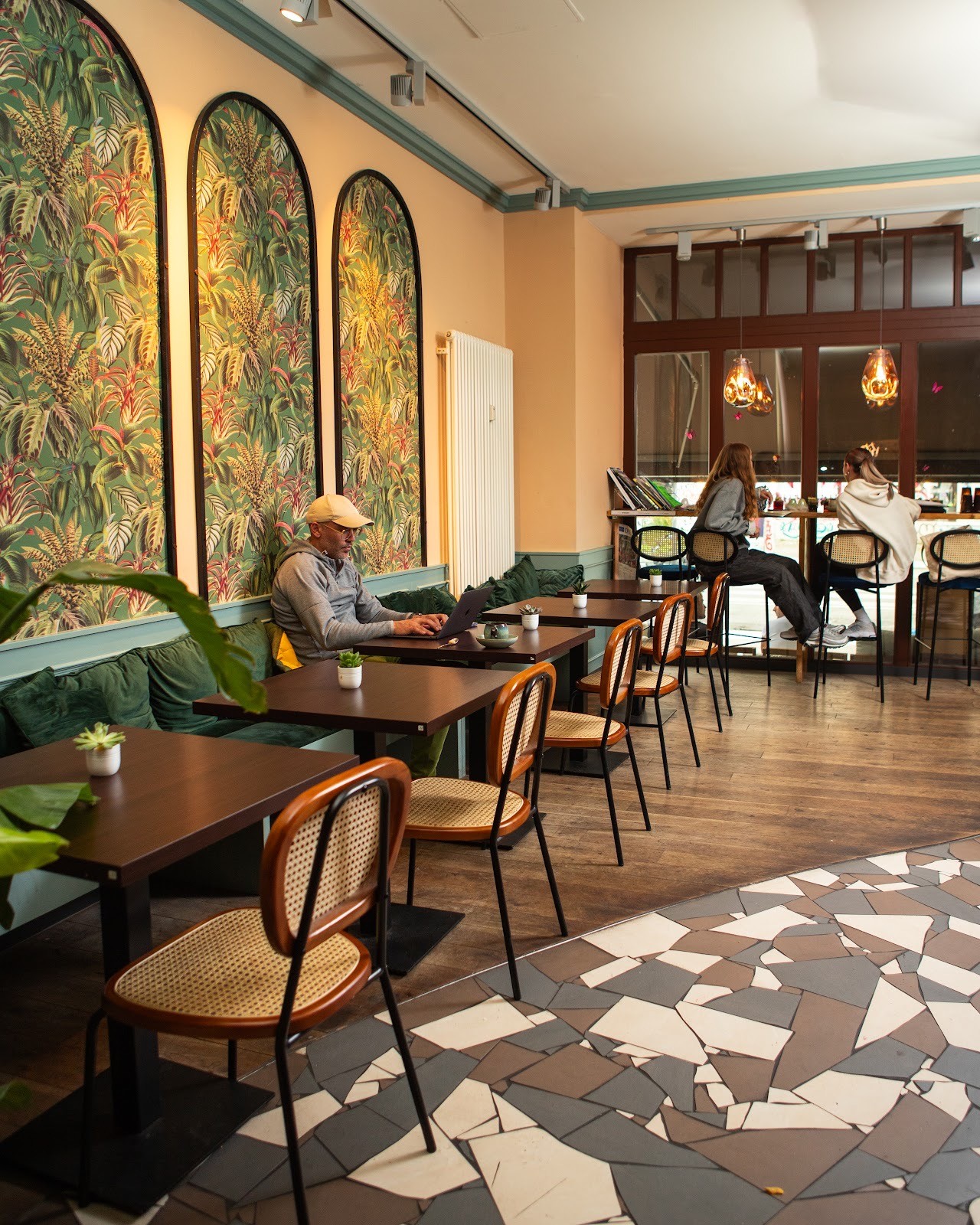 Interior shot of Jederzeit Cafe with a man working on a laptop at a table, and other patrons in the background, featuring tropical wallpaper and wooden floors.