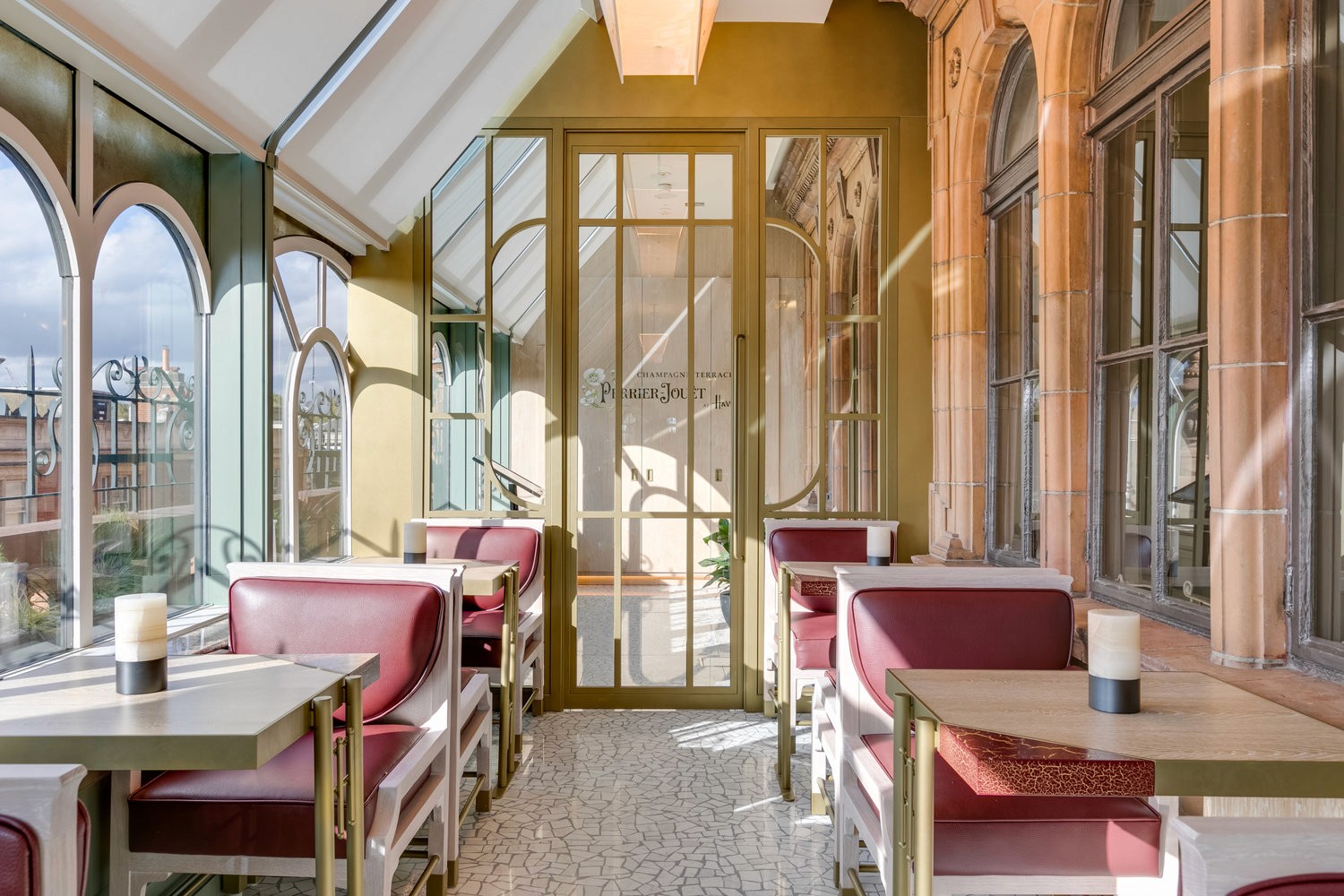 A sunlit café with arched windows, red leather chairs, and small wooden tables set against elegant architecture.