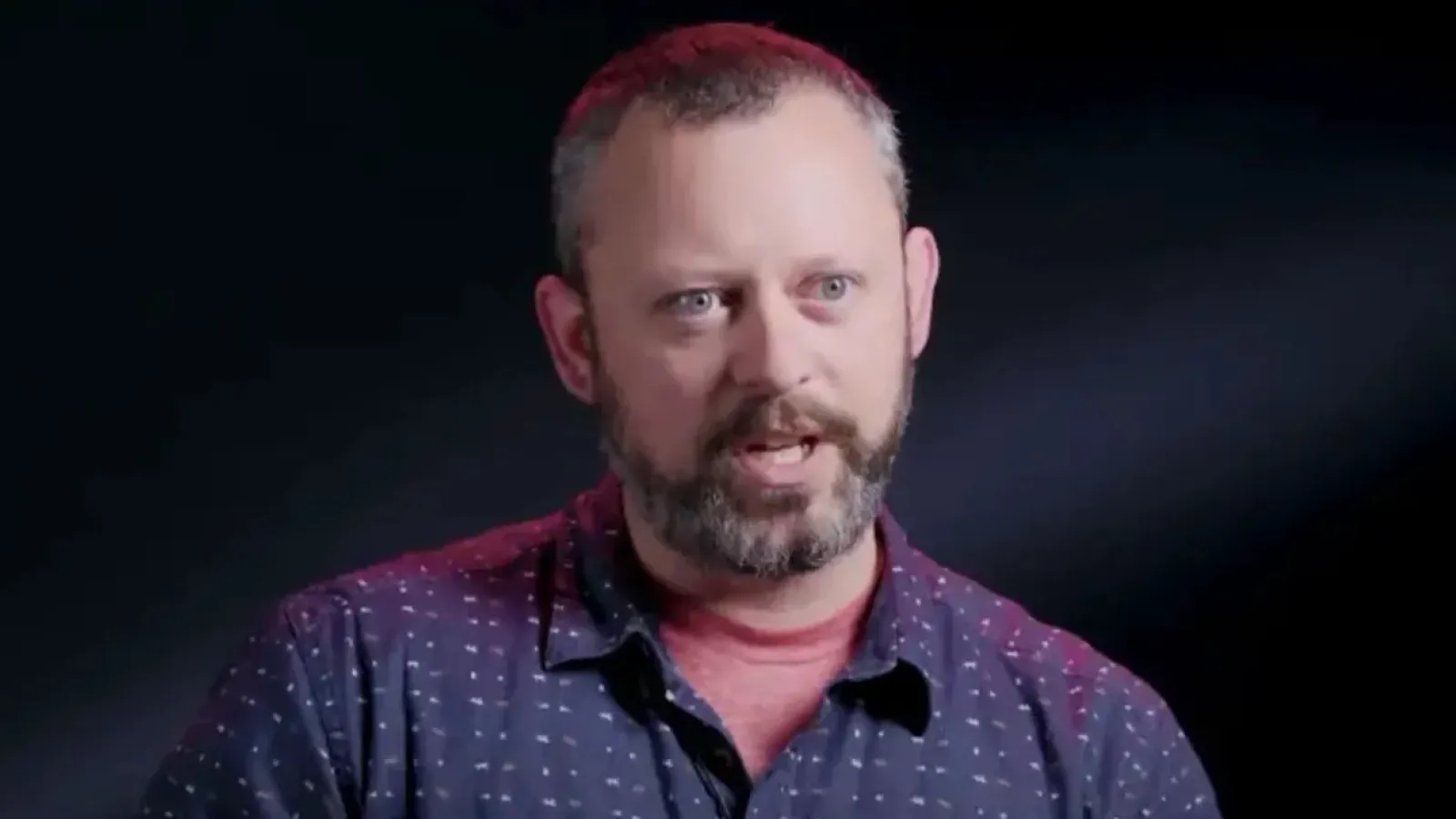 A man with a short beard and blue eyes is speaking, wearing a navy blue button-up shirt with small white patterns, against a dark, softly lit background.