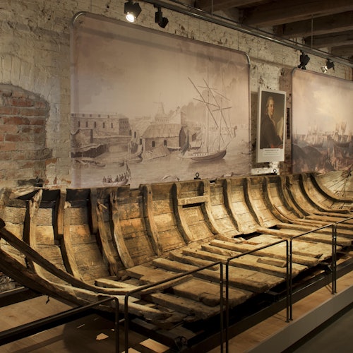 Museossa esillä oleva puisen laivan runko, taustaseinällä historiallisia merenkulun maalauksia ja muotokuvia.