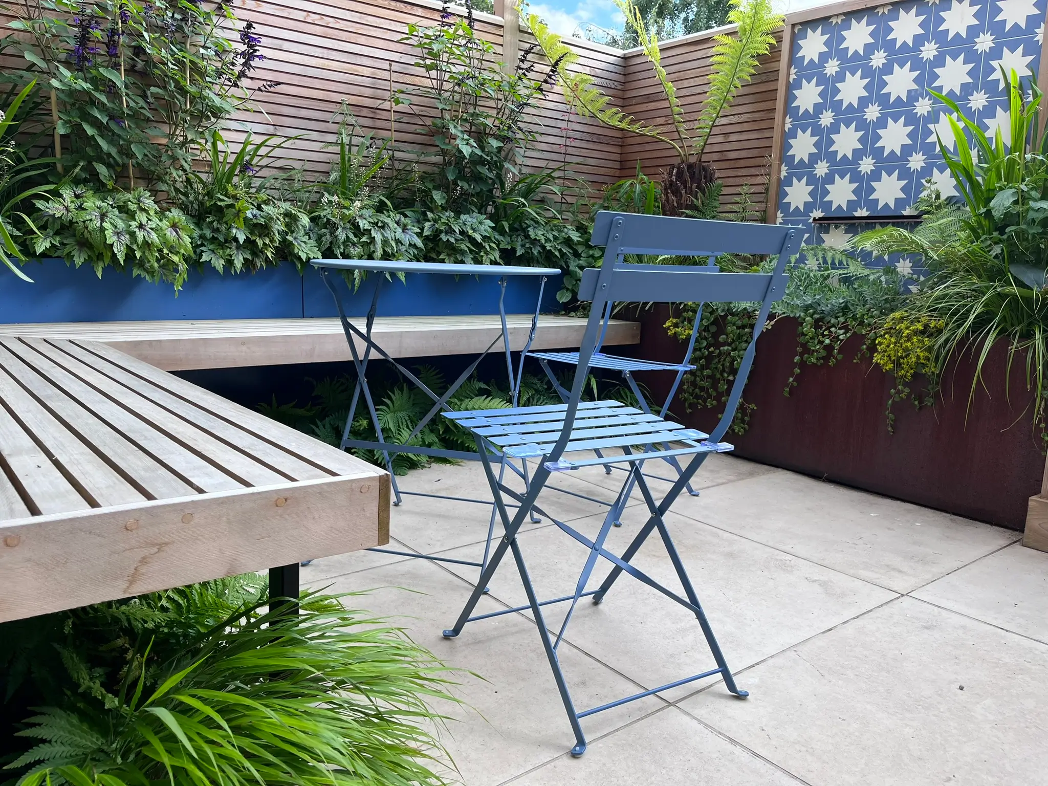 A small outdoor patio with a blue folding table and chairs surrounded by greenery and a wooden deck.