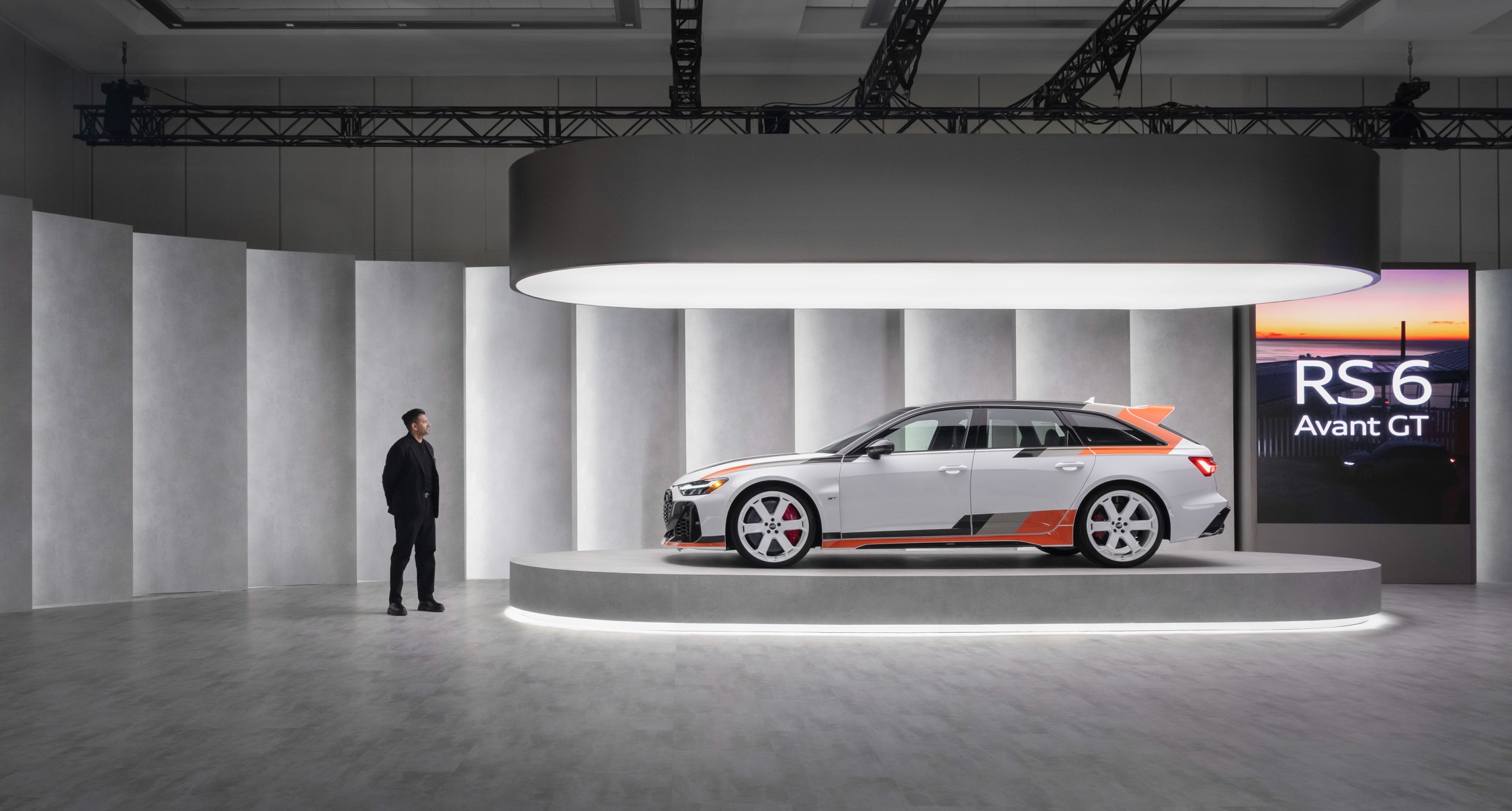 A sleek white Audi RS 6 Avant GT with orange accents is displayed on a modern, illuminated stage. A person in black observes it, highlighting luxury and innovation.