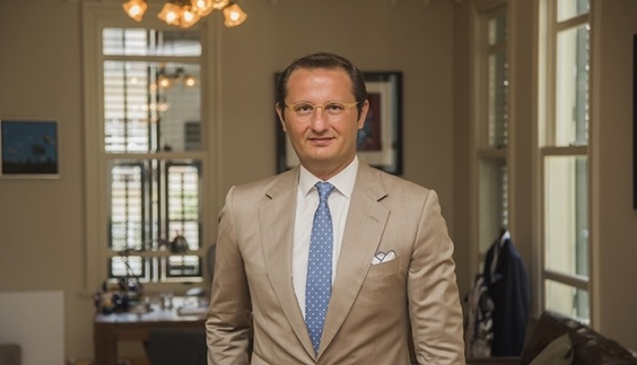 Man in a blue t-shirt smiling confidently while standing against a light wall in sunlight
