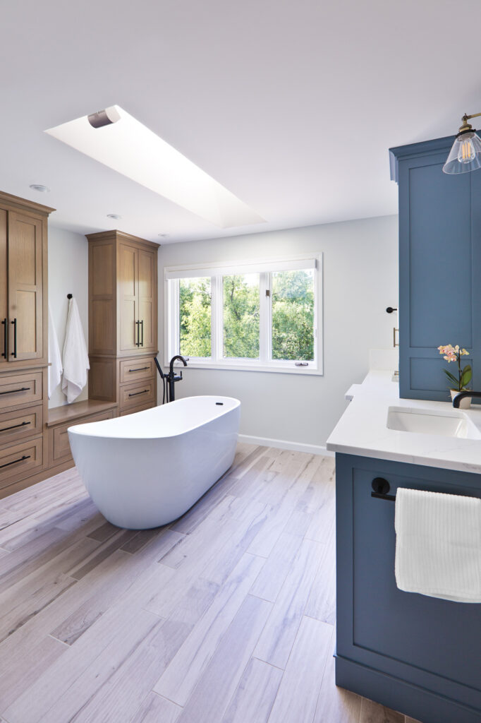 Bright modern bathroom featuring a white freestanding tub, light wood floors, large skylight, and built-in wooden and blue cabinetry. A walk-in glass shower with black fixtures sits near a private toilet room.