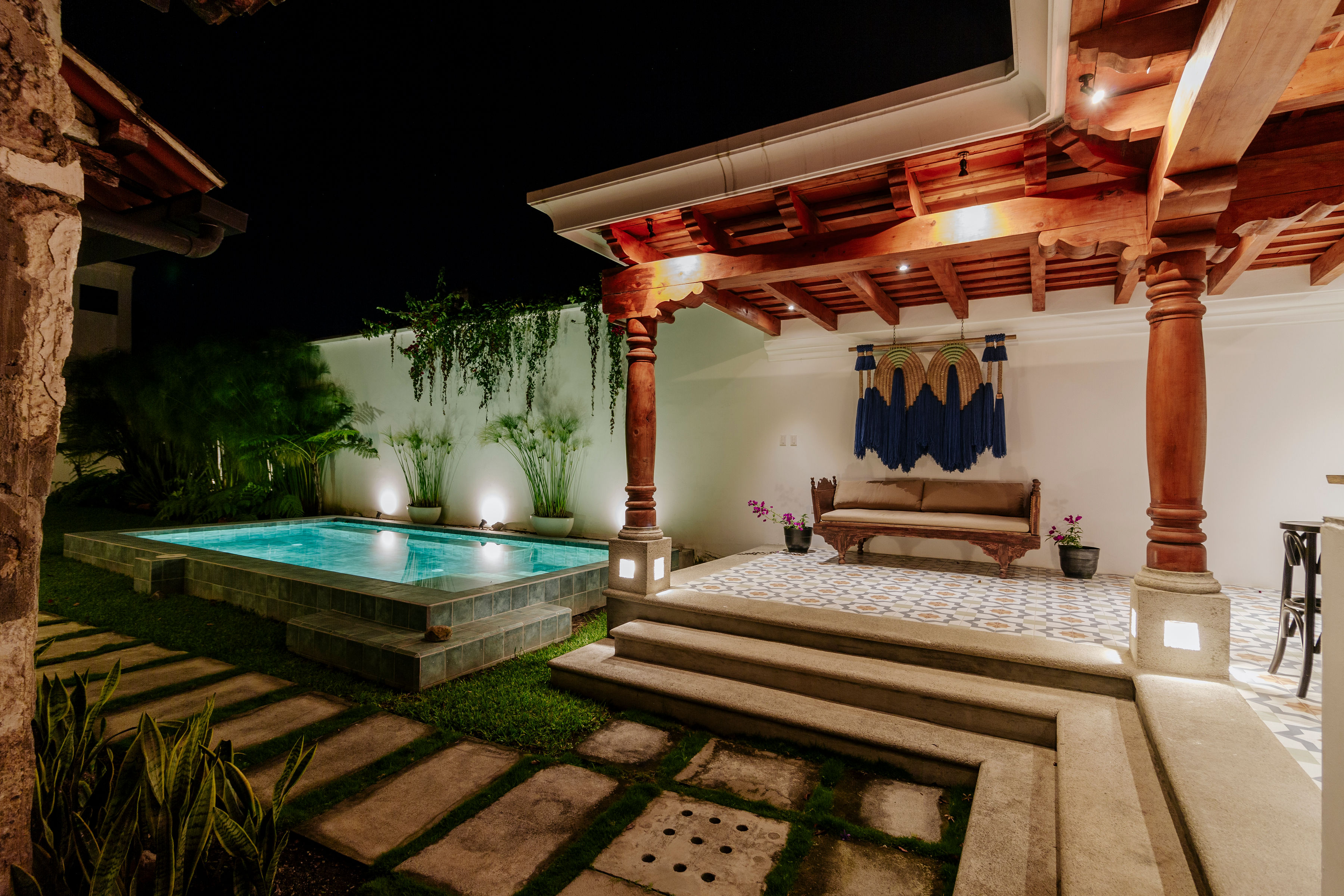 A beautifully lit outdoor patio at night features a cozy seating area under a wooden pergola, adjacent to a modern rectangular swimming pool surrounded by lush greenery.