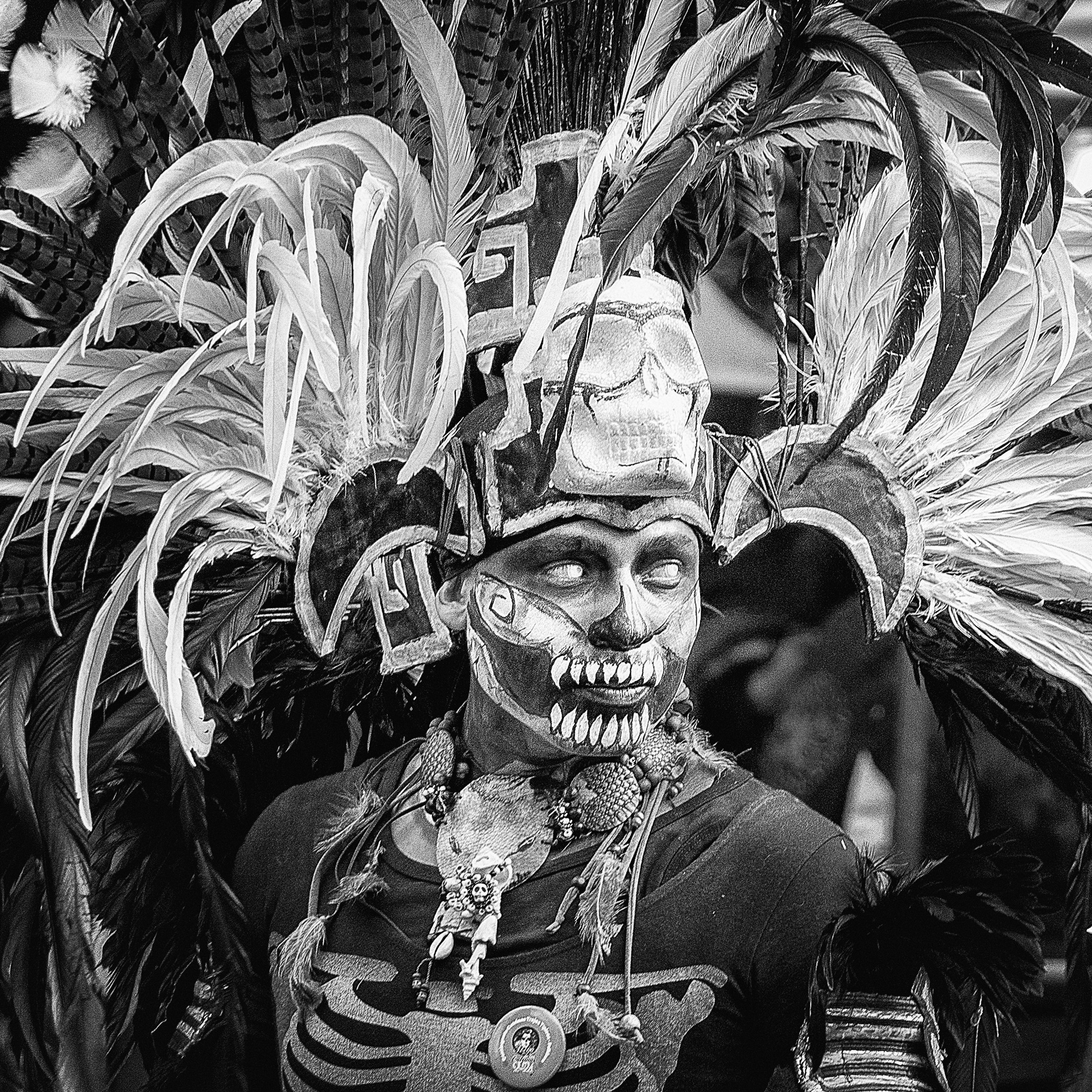 Black and white portrait of a man in elaborate Day of the Dead ceremonial costume, featuring face paint, skull motifs, feathered headdress, and bone-like chest markings.