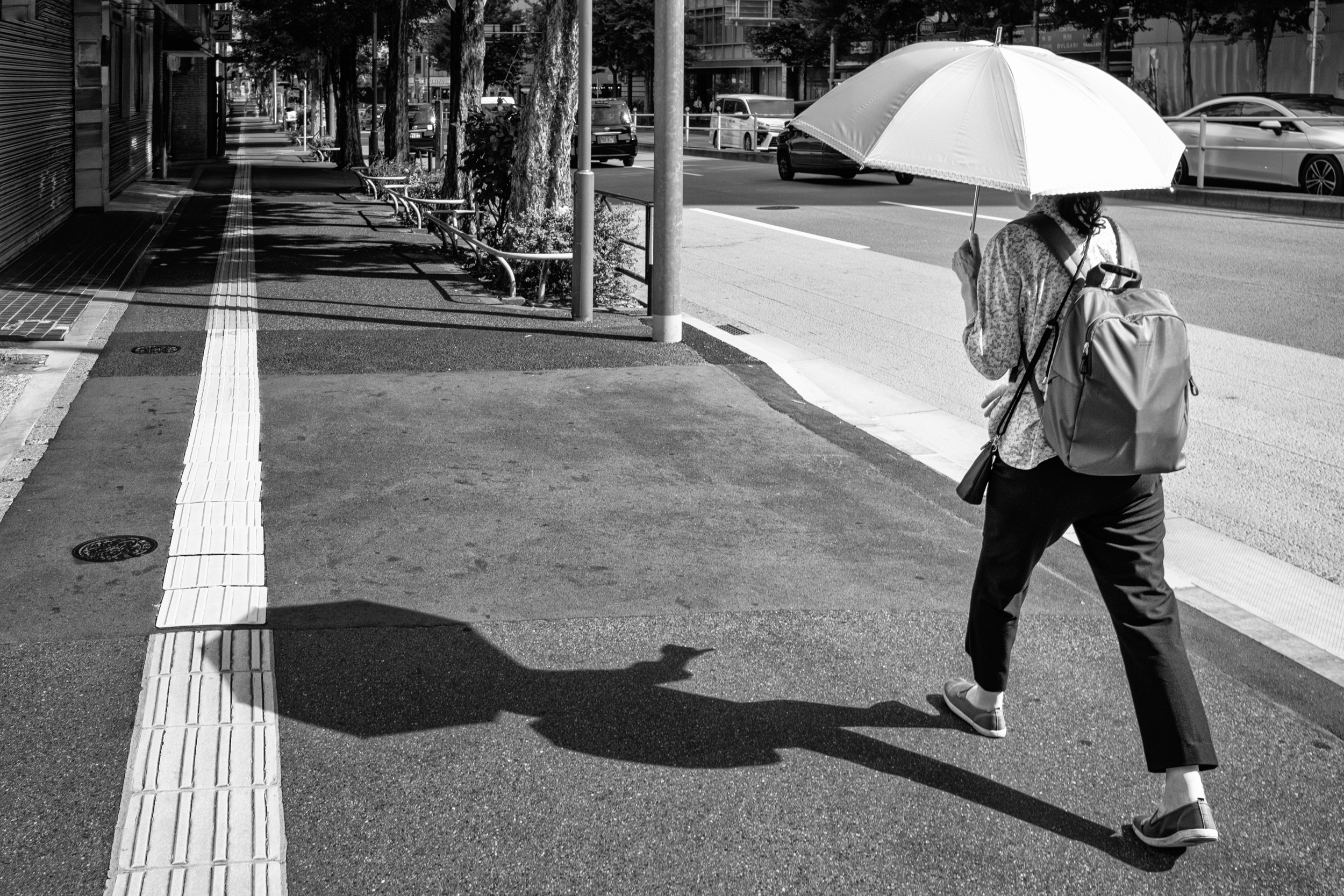 Black and white street scene of a person walking on a sidewalk holding a backpack and a large umbrella.