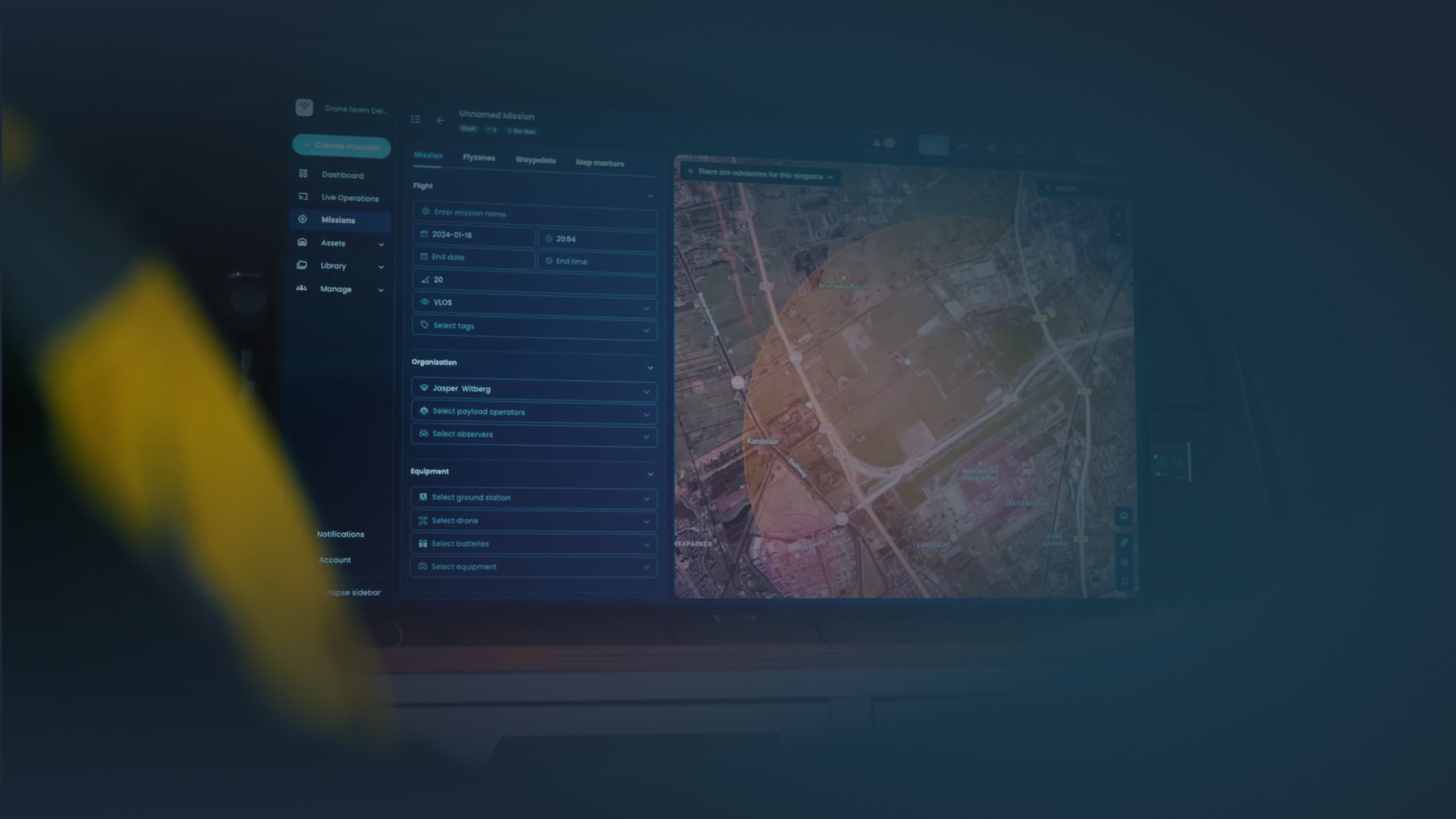 Ein großer Monitor zeigt die AirHub-Drohnenverwaltungsplattform in einem dunklen, operativen Umfeld. Auf dem Bildschirm ist eine detaillierte Live-Karte mit Flugzonen und Telemetriedaten zu sehen, begleitet von einer Missionsleiste mit Flugparametern und Geräteeinstellungen. Im Vordergrund deutet die verschwommene Schulter einer Person in einer hochsichtbaren Uniform auf ein Kommandozentrum oder eine mobile Einheit hin.